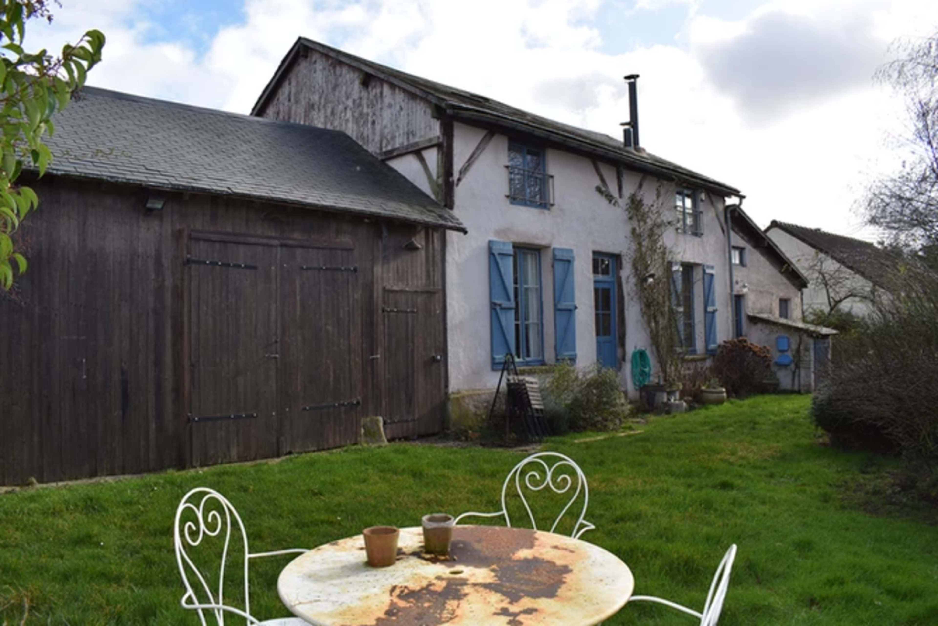 A two-story house with blue shutters and a large wooden barn is set in a grassy yard featuring a rusted metal table and two white chairs.