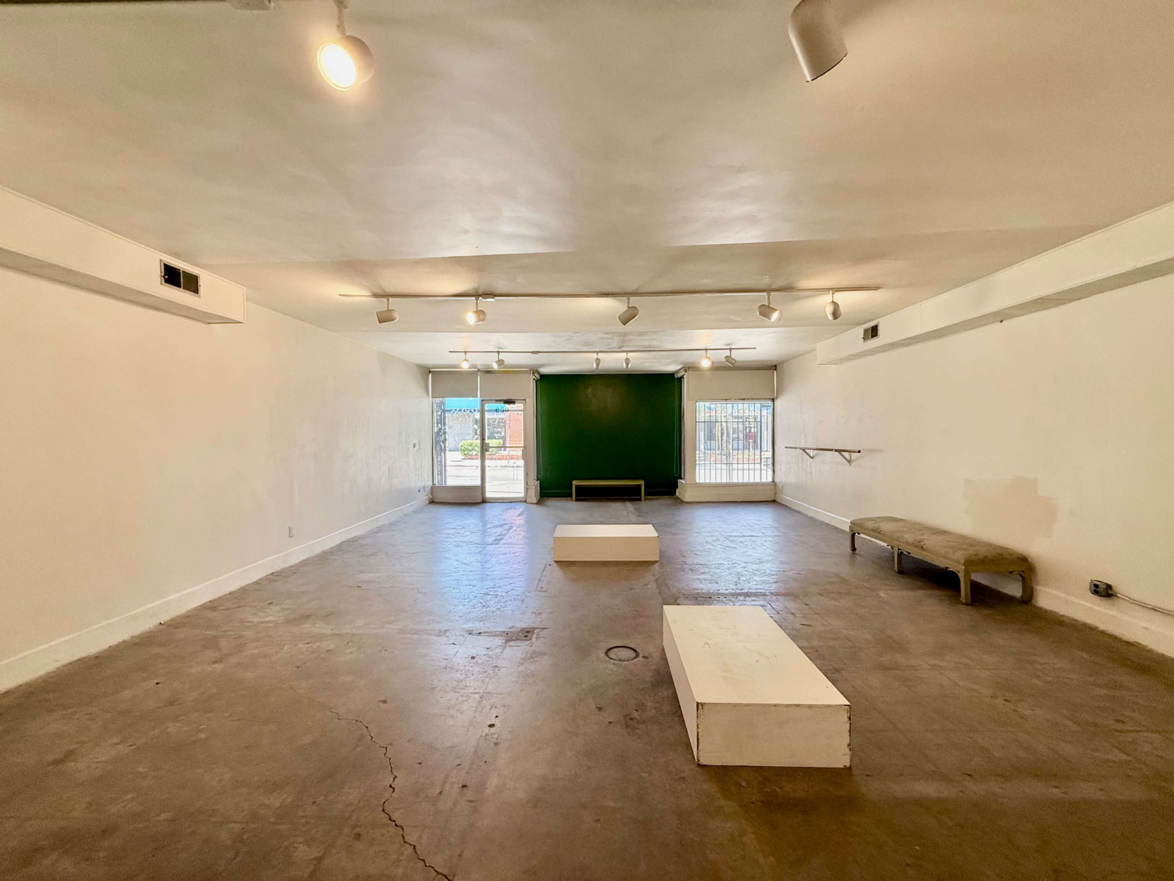 The image shows a spacious, unfurnished room with white walls, a green backdrop at one end, and a couple of simple white platforms in the center.