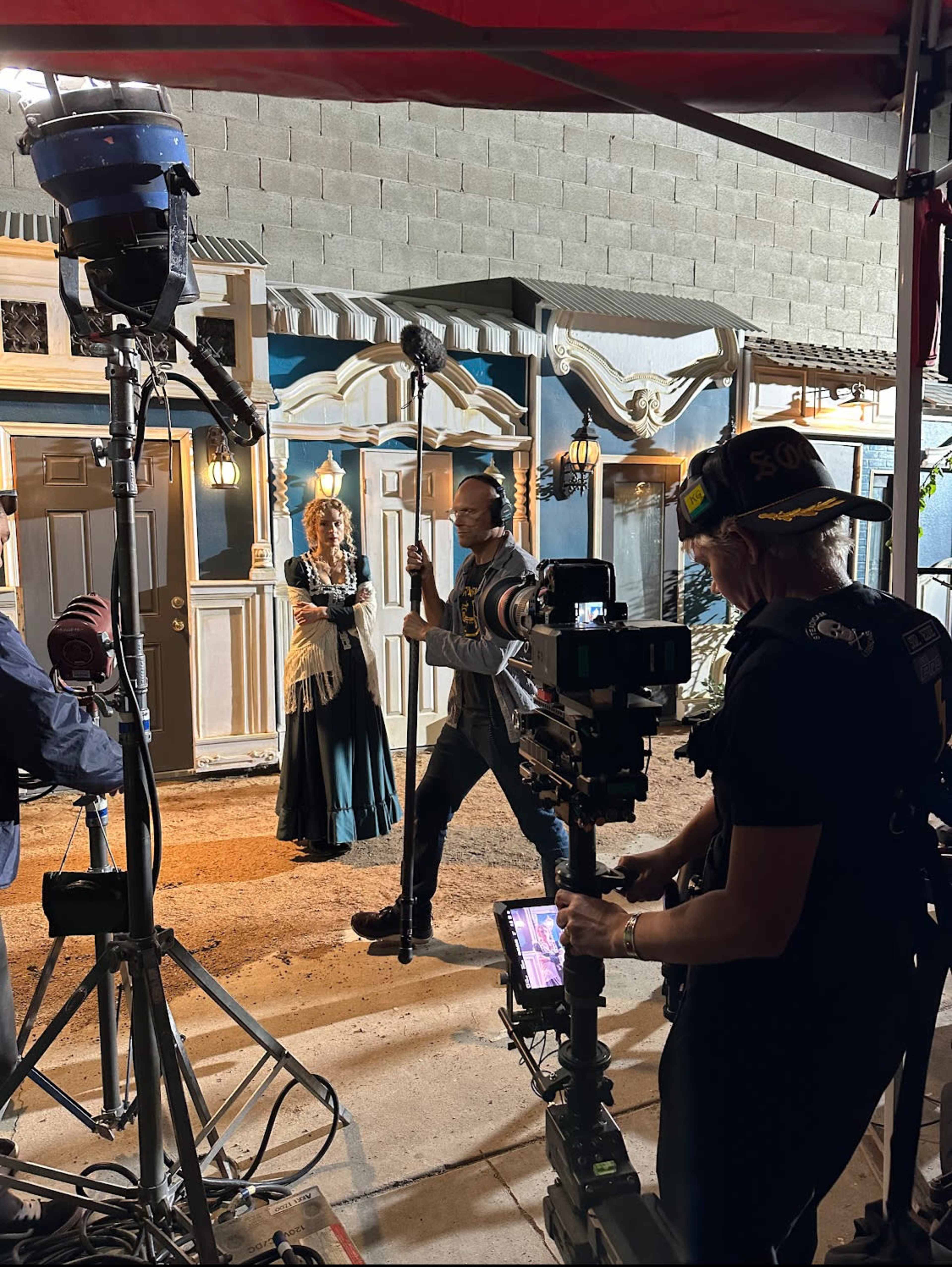 A film crew is set up outside a building, with a woman in period costume and a man gesturing as they prepare to shoot a scene.