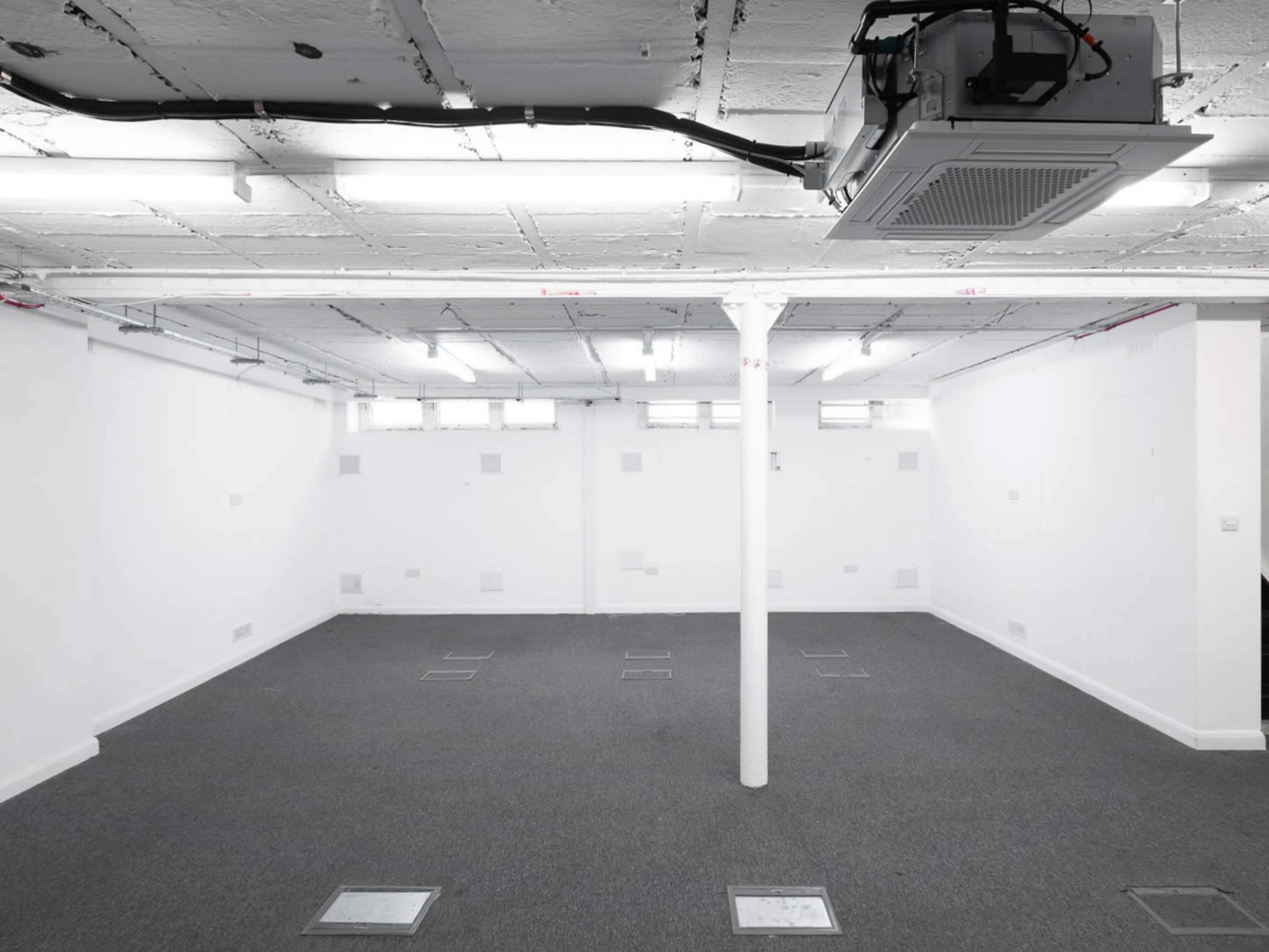 The image shows a sparsely furnished room with gray carpet, white walls, and a central support pillar, along with ceiling-mounted lighting and air conditioning.