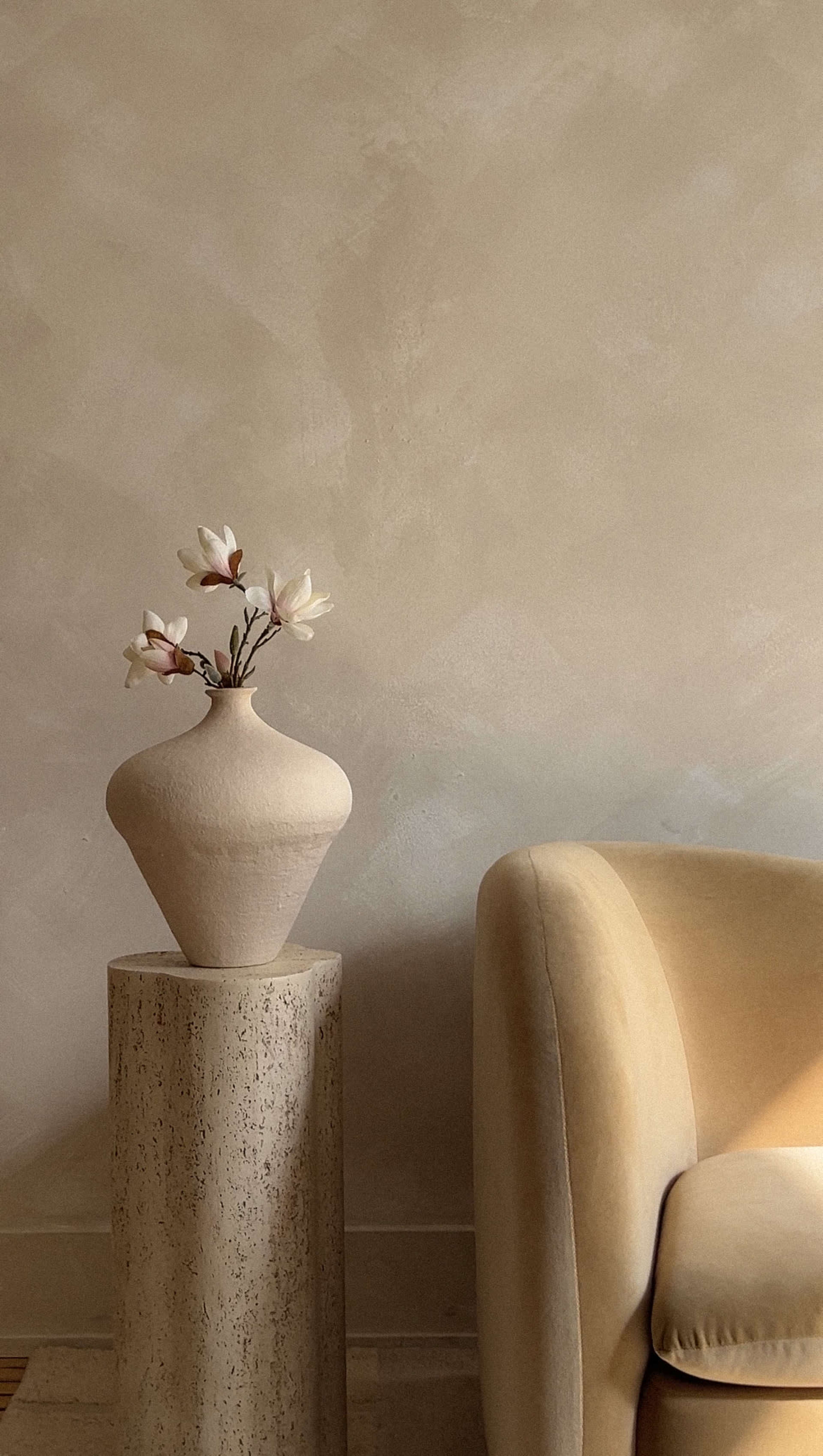 A light-colored vase with flowers sits on a textured stone pedestal next to a beige upholstered chair against a softly colored wall.
