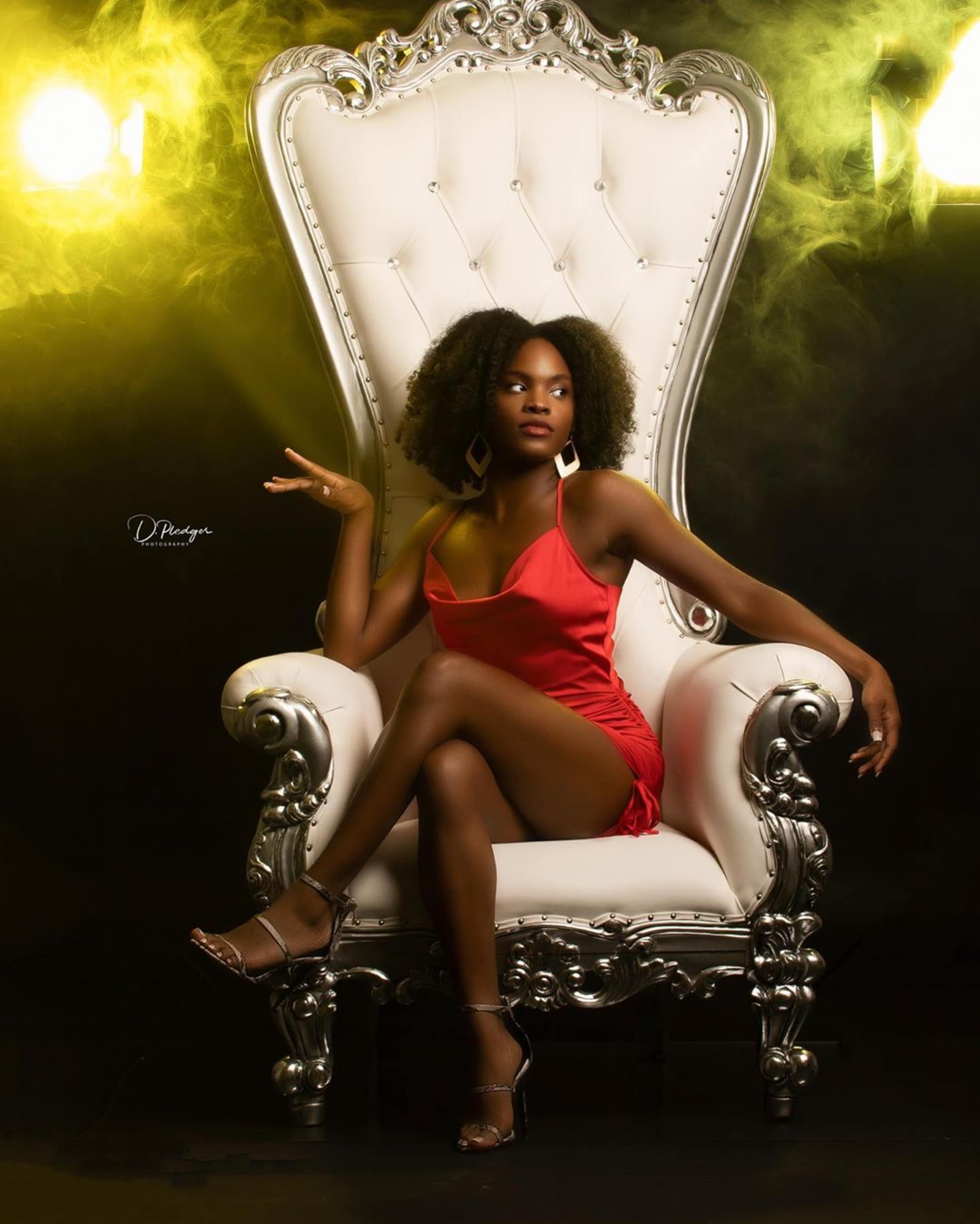 A woman in a red dress sits elegantly on a large ornate white chair against a dark backdrop with dramatic lighting.