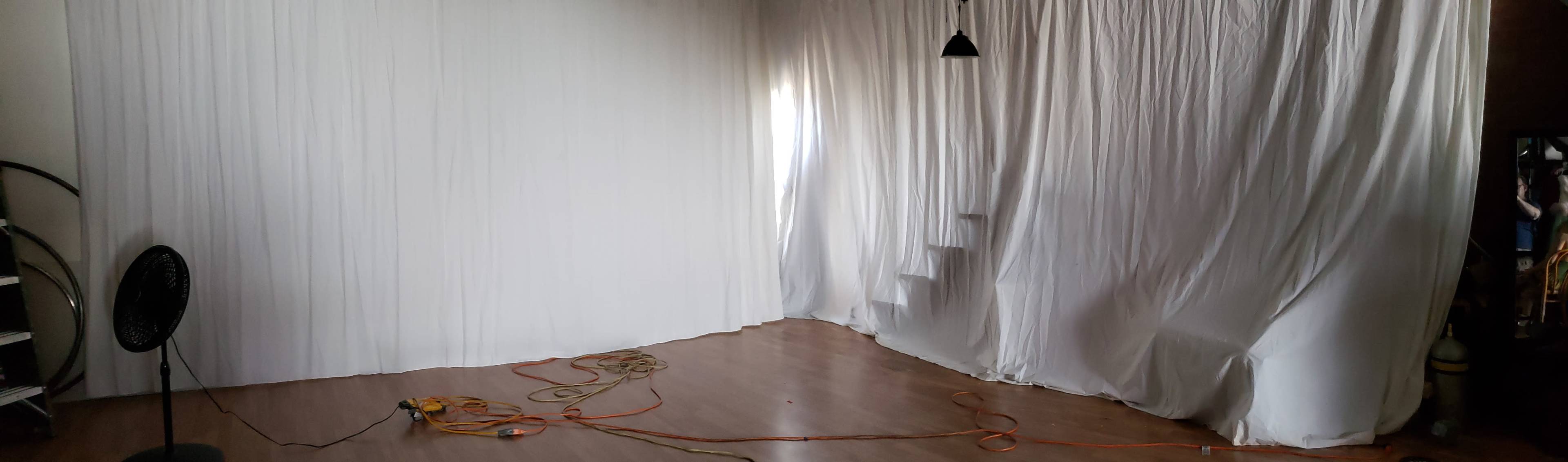 A large, white-draped room with a wooden floor, a fan, and a visible staircase behind the drapery.