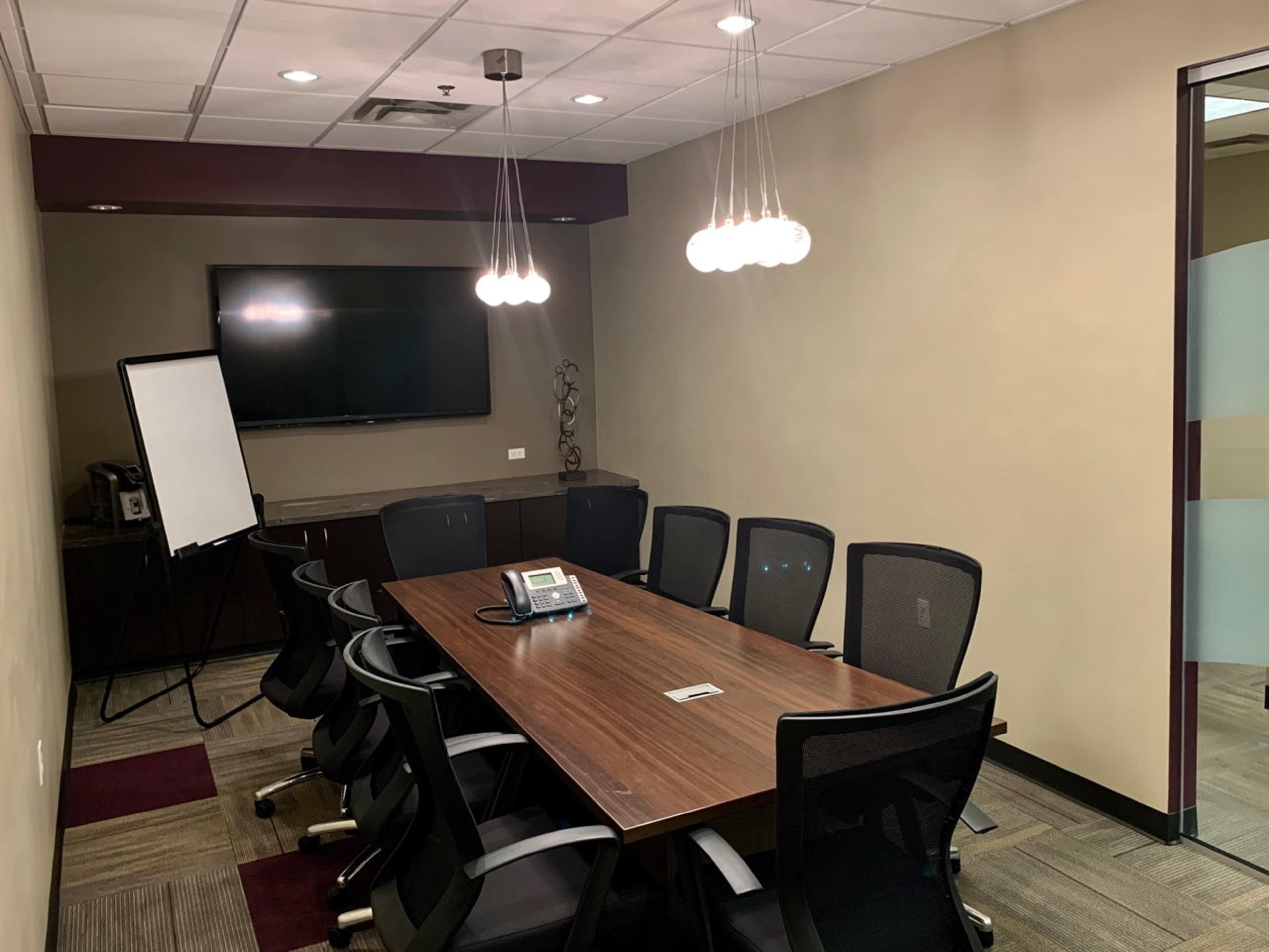 A conference room features a long wooden table surrounded by black mesh chairs, with a television mounted on the wall and a whiteboard in the corner.