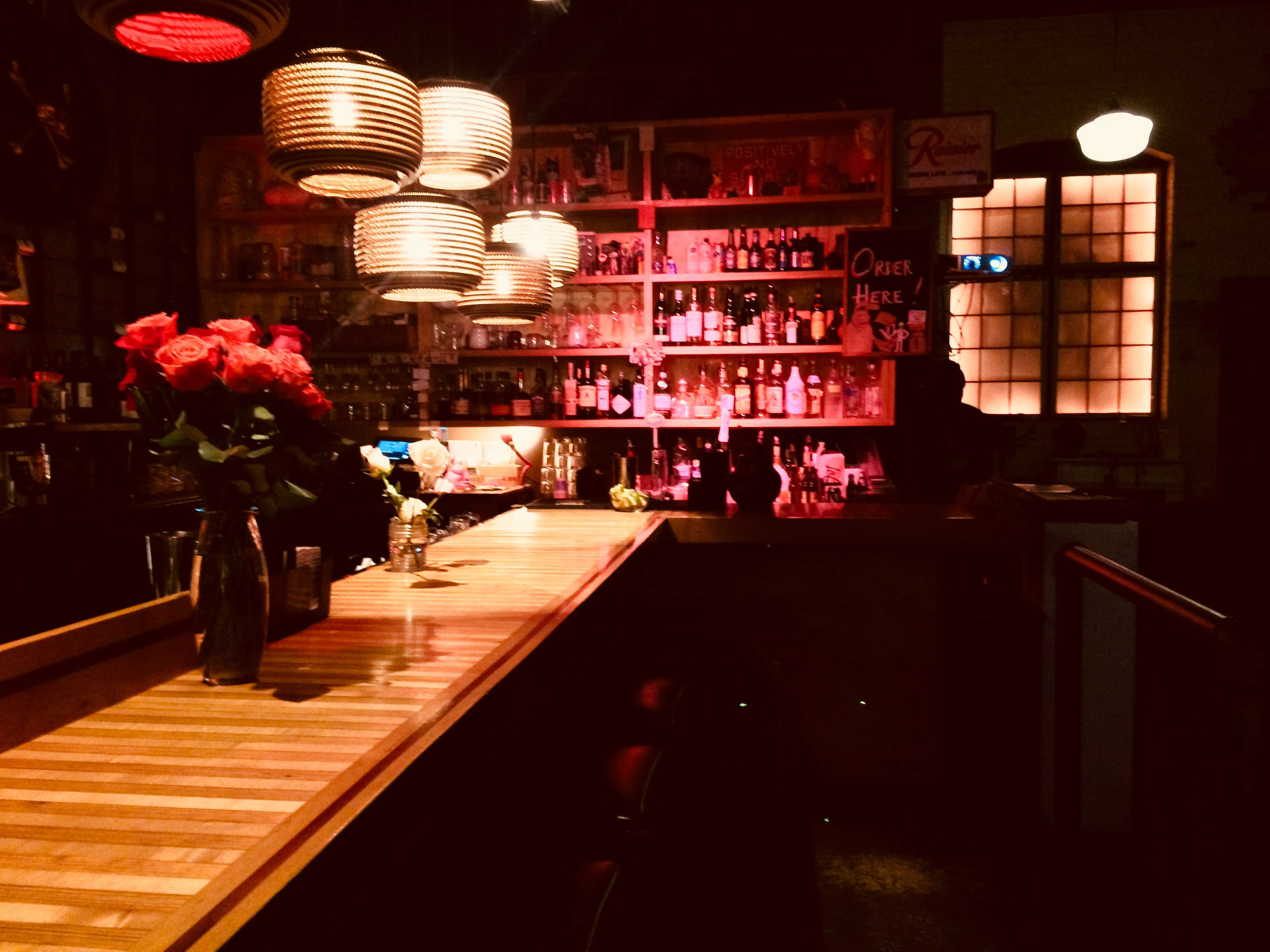 A dimly lit bar with a wooden counter, shelves lined with various bottles, and a vase of roses on the bar.