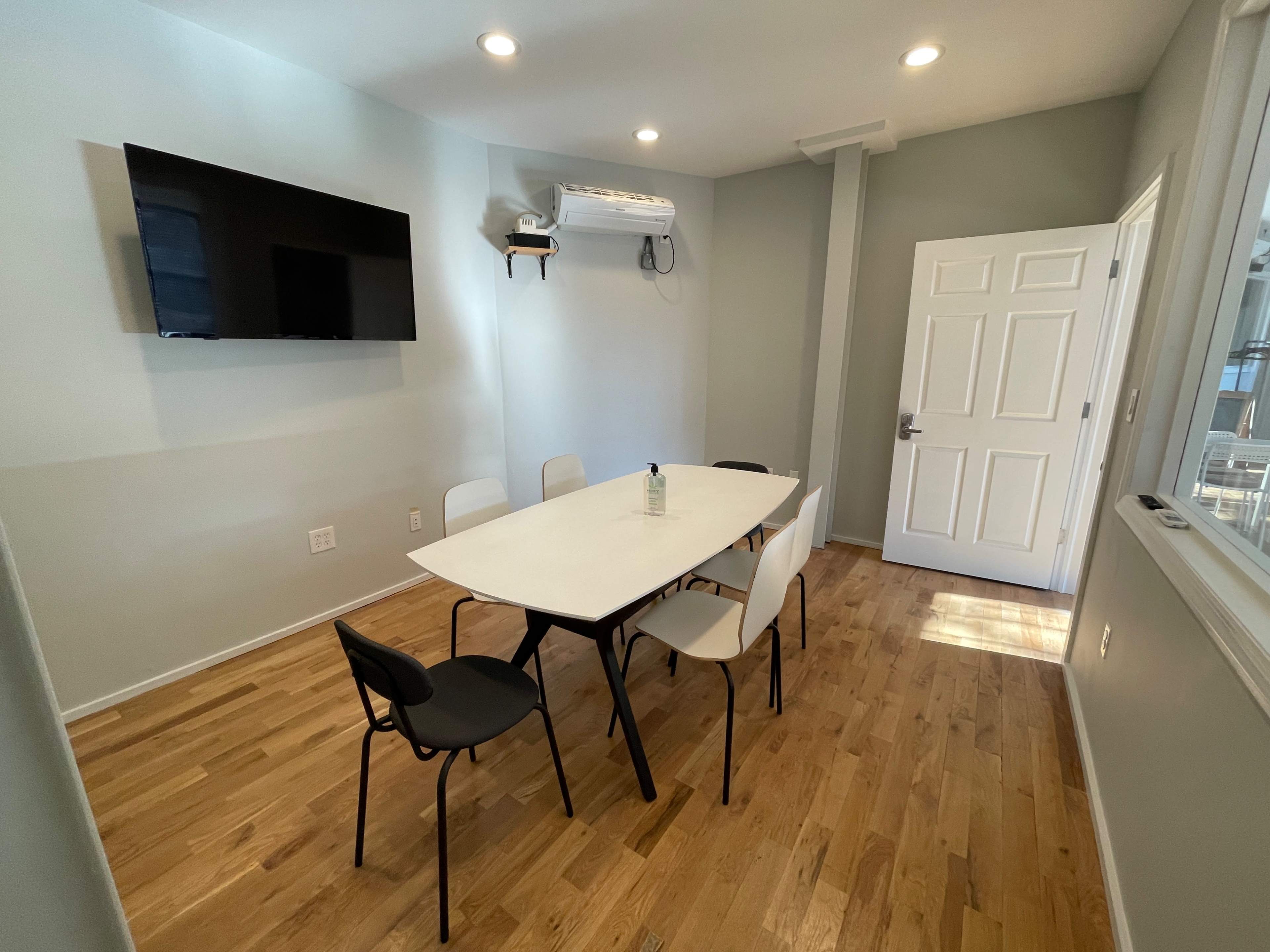 A meeting room features a rectangular table surrounded by six chairs, with a television mounted on the wall and an air conditioning unit above.