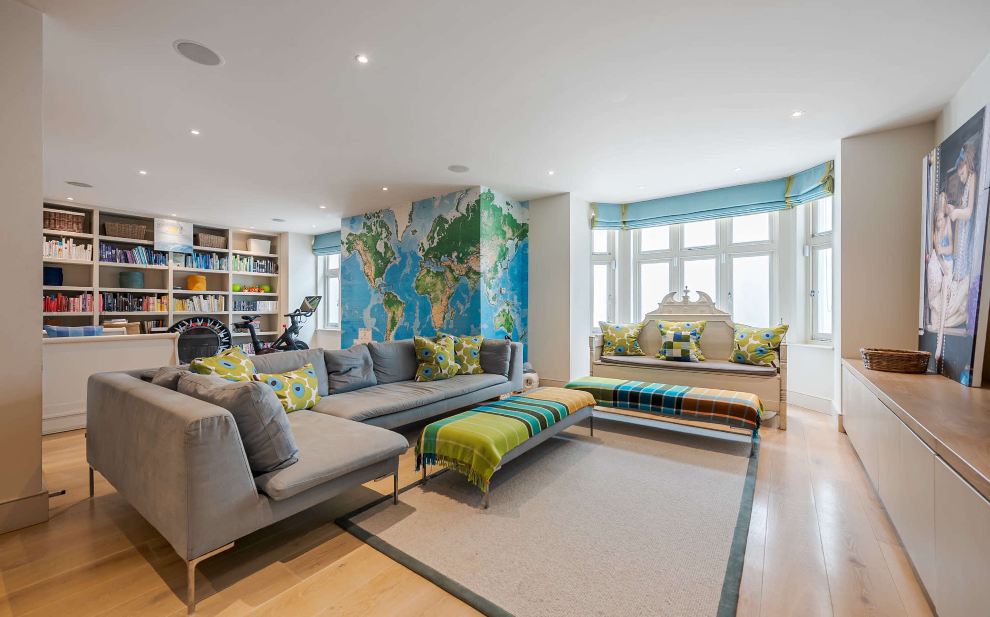 A modern living room features a large world map mural, a gray sofa with colorful cushions, and natural light from large windows.
