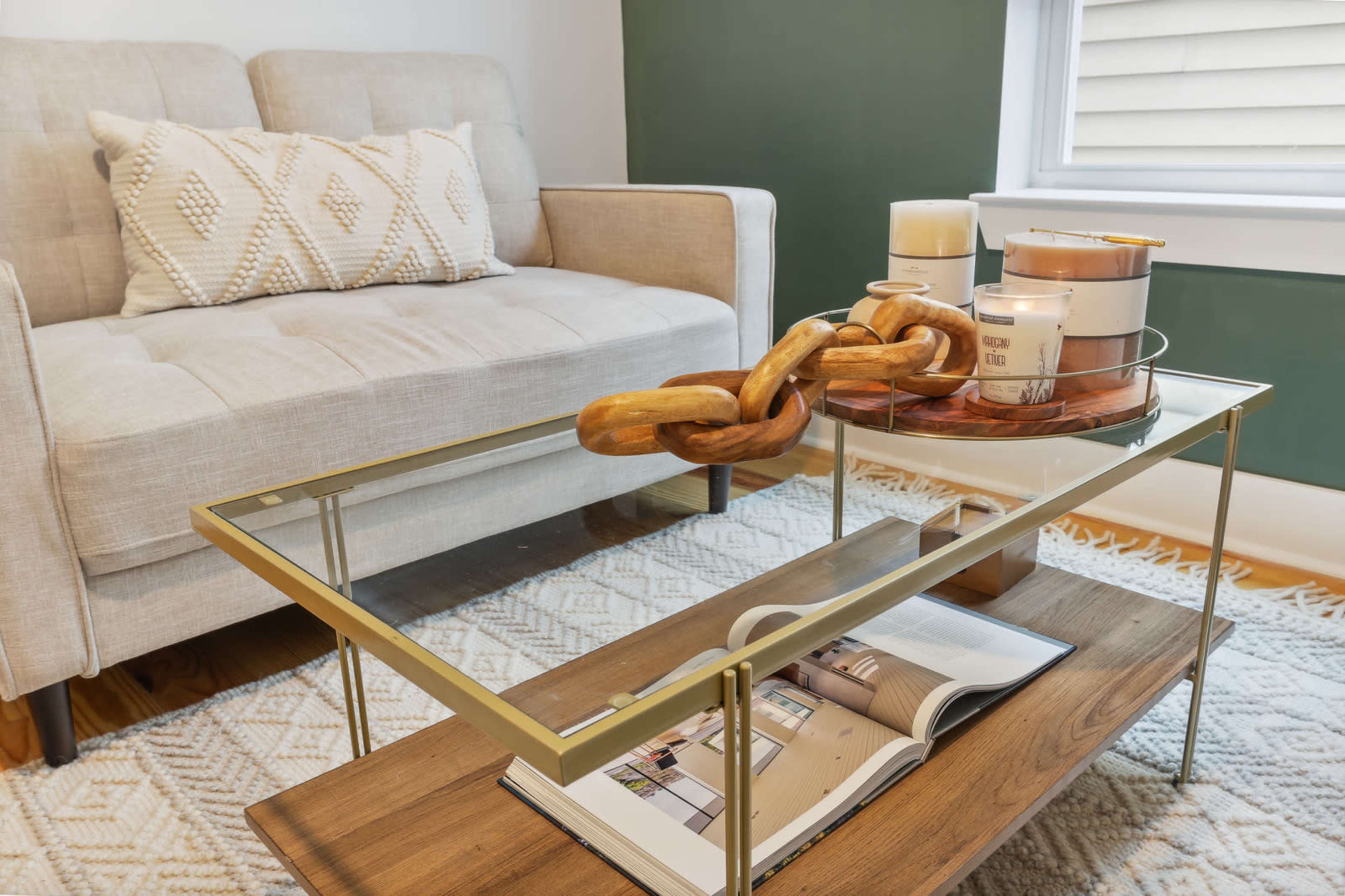 A modern living room scene features a beige sofa with decorative cushions, a glass and gold coffee table holding a wooden chain sculpture and candles, and a magazine on a wooden shelf below.