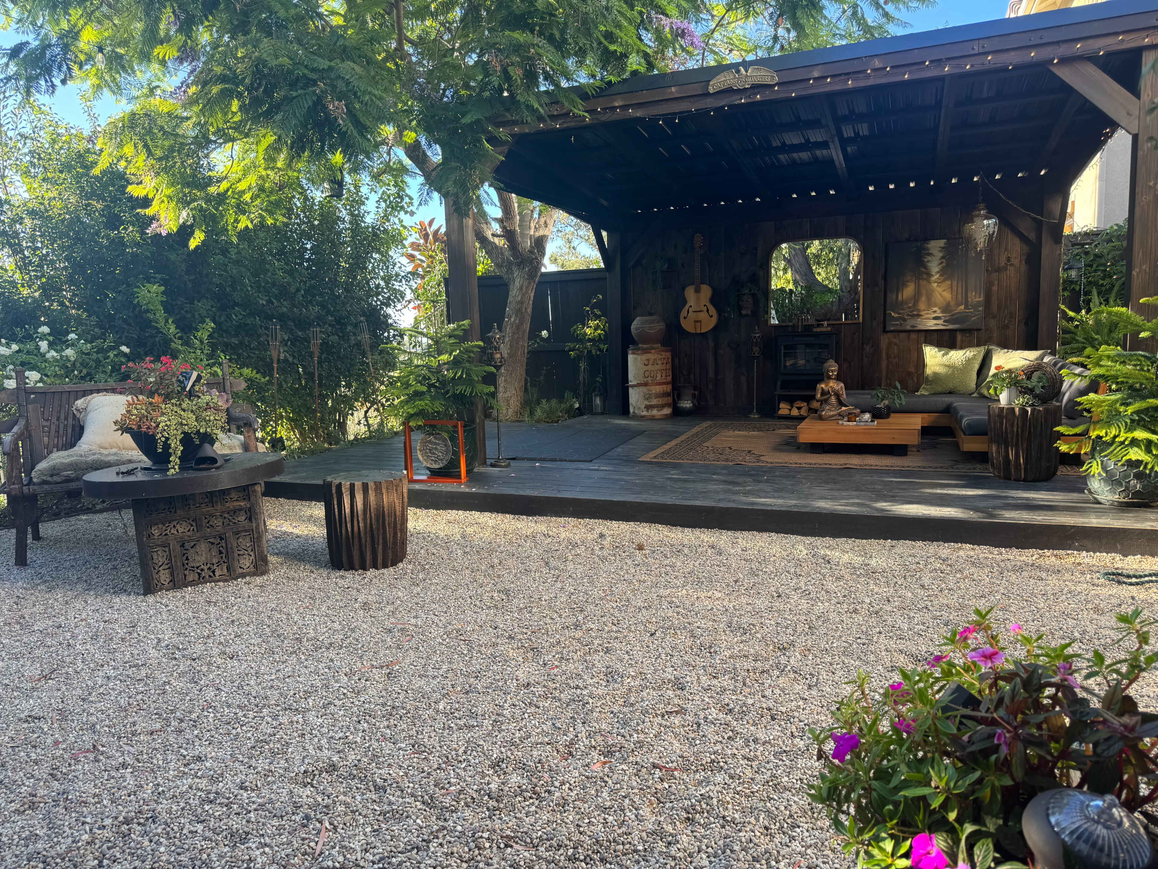 The image shows a landscaped outdoor space featuring a wooden gazebo with seating, decorative plants, a gravel surface, and various furnishings.