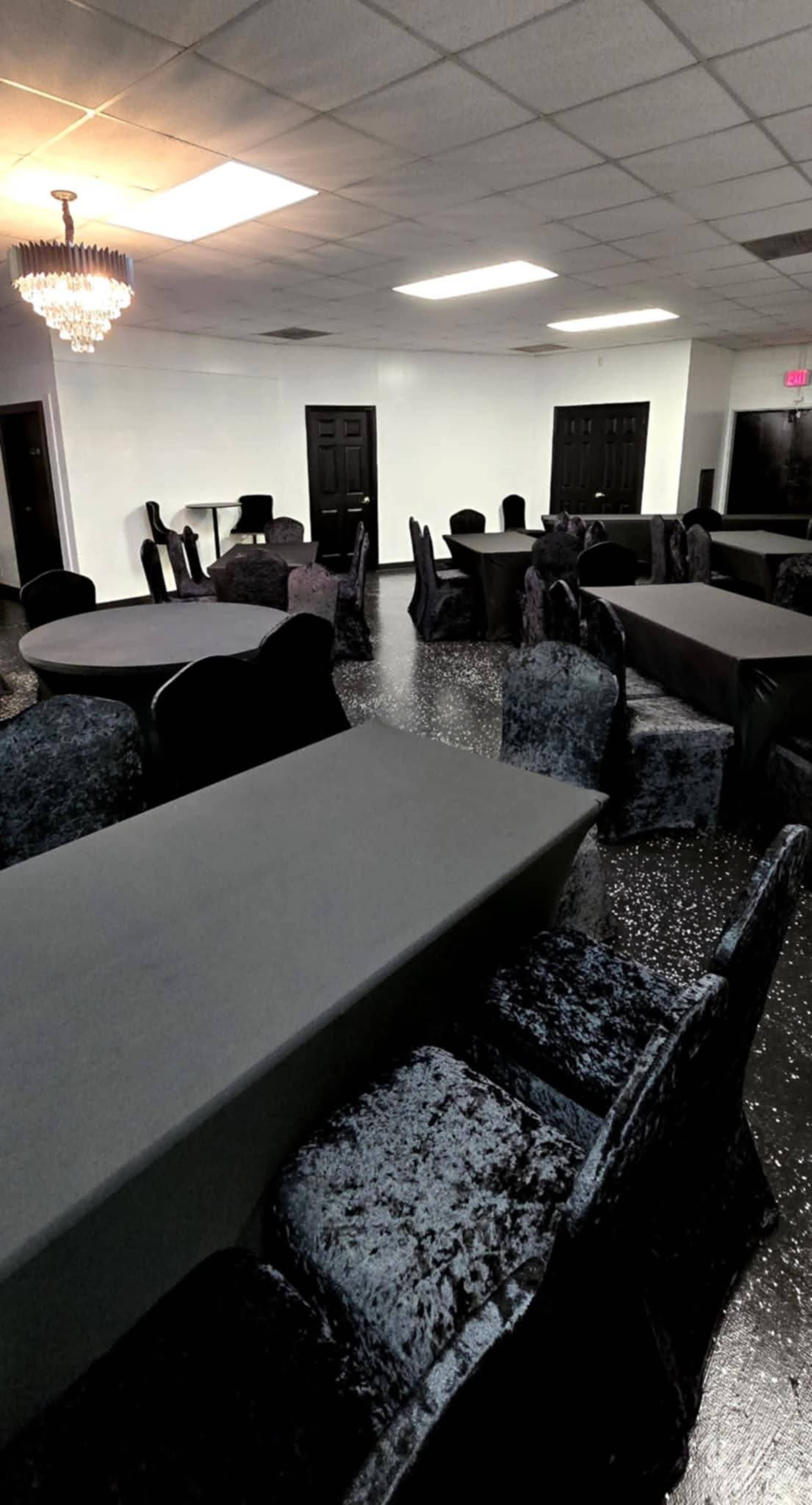 The image shows a dimly lit event space with multiple tables covered in black tablecloths, surrounded by black upholstered chairs.