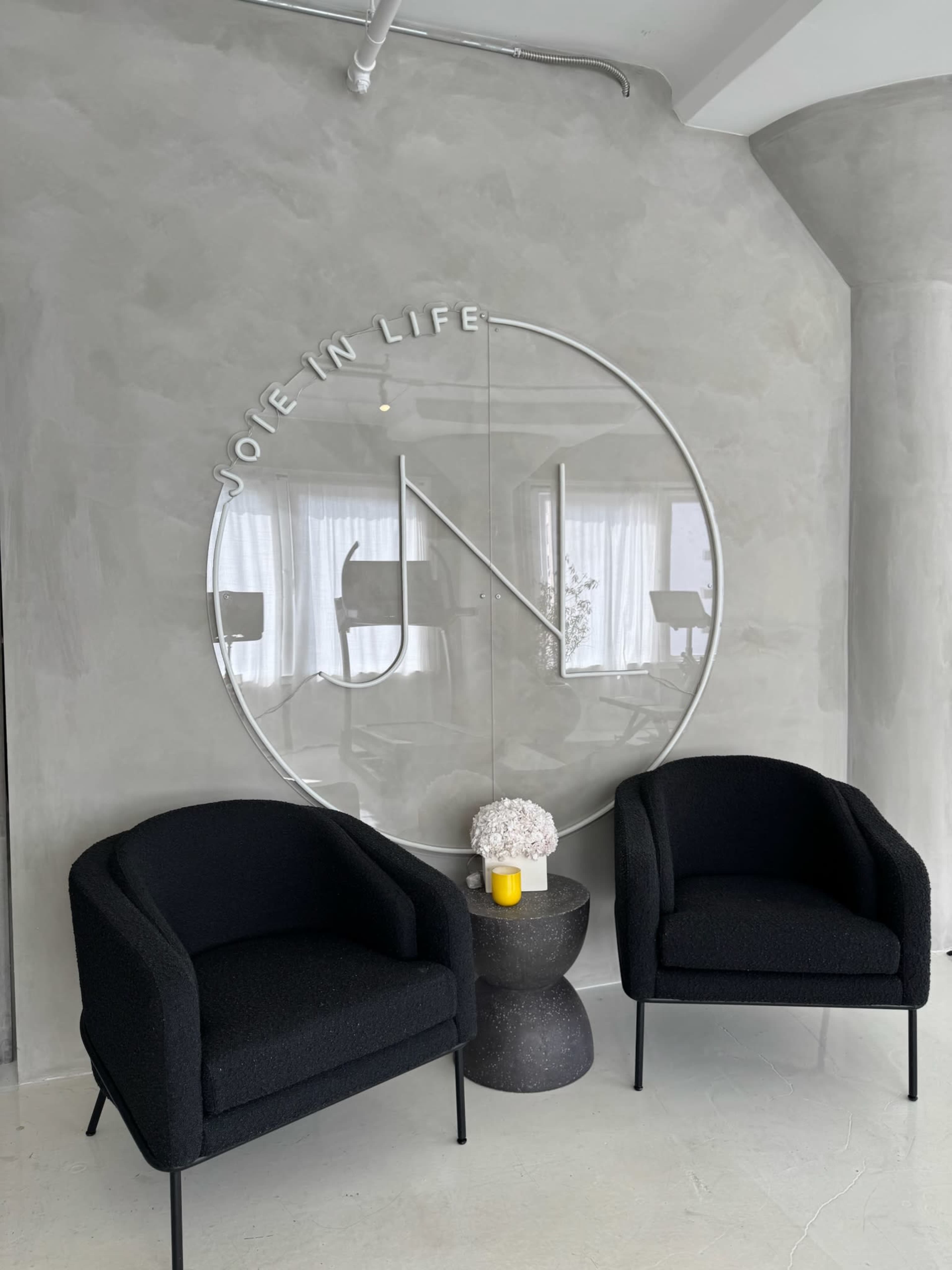 The image shows a minimalist indoor space featuring two black chairs facing a round mirror with the phrase "JOIE IN LIFE" etched into it, along with a small table and a vase of flowers.