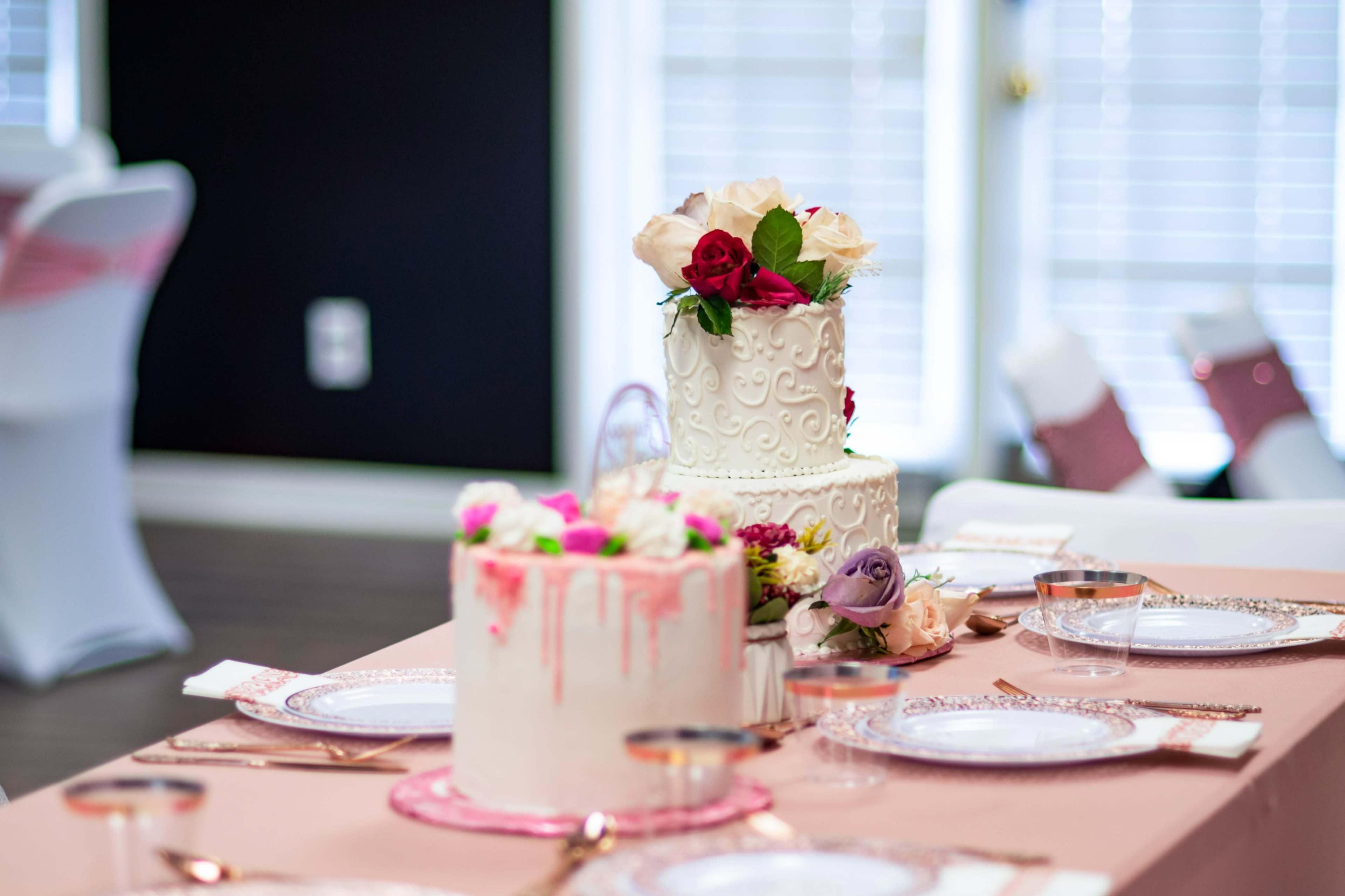 Two elegantly designed wedding cakes with decorative flowers sit on a table set with fine dishware and a pink tablecloth.