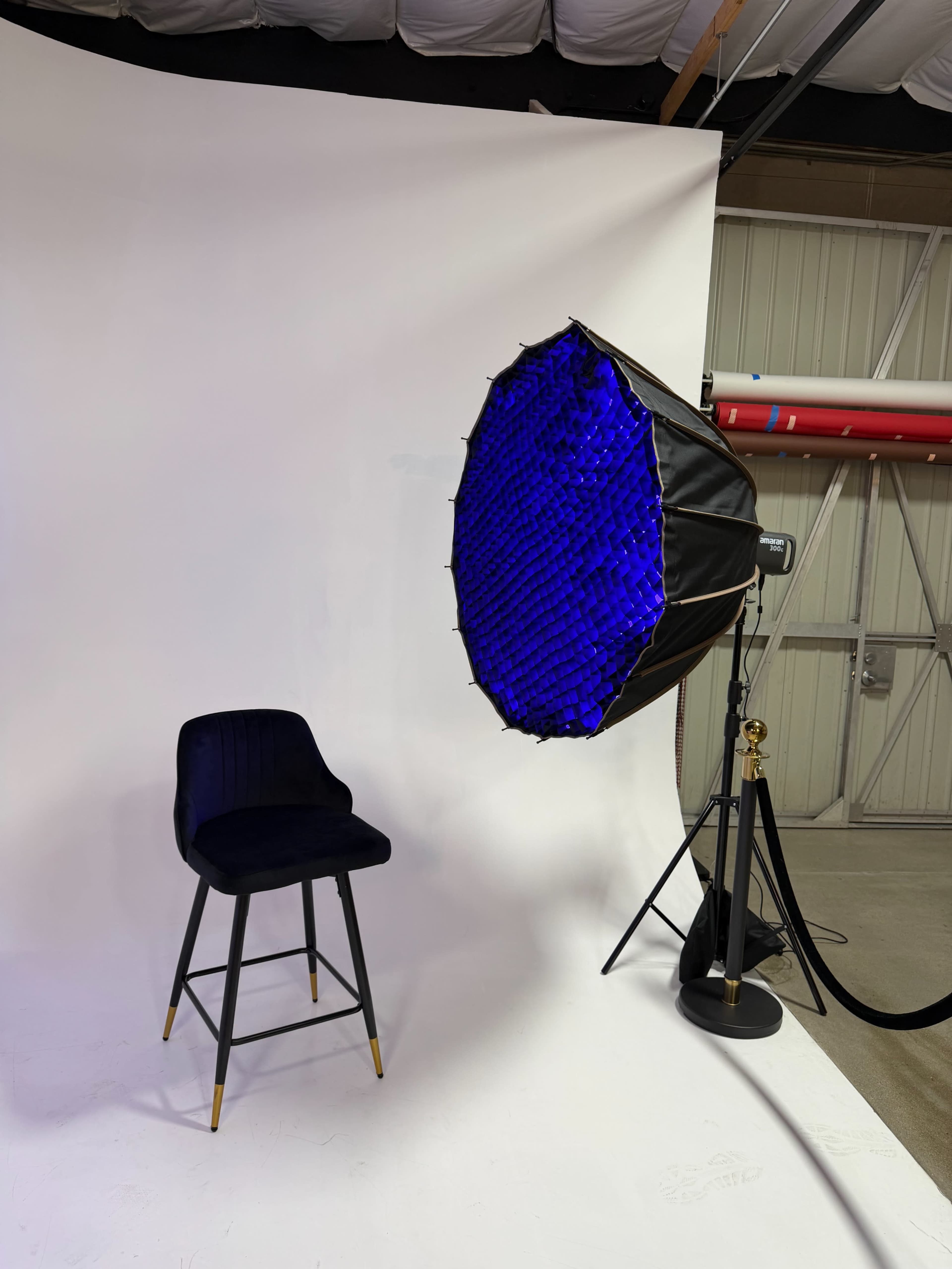 The image shows a studio setup with a tall black stool beside a large softbox light with a blue interior, against a white backdrop.