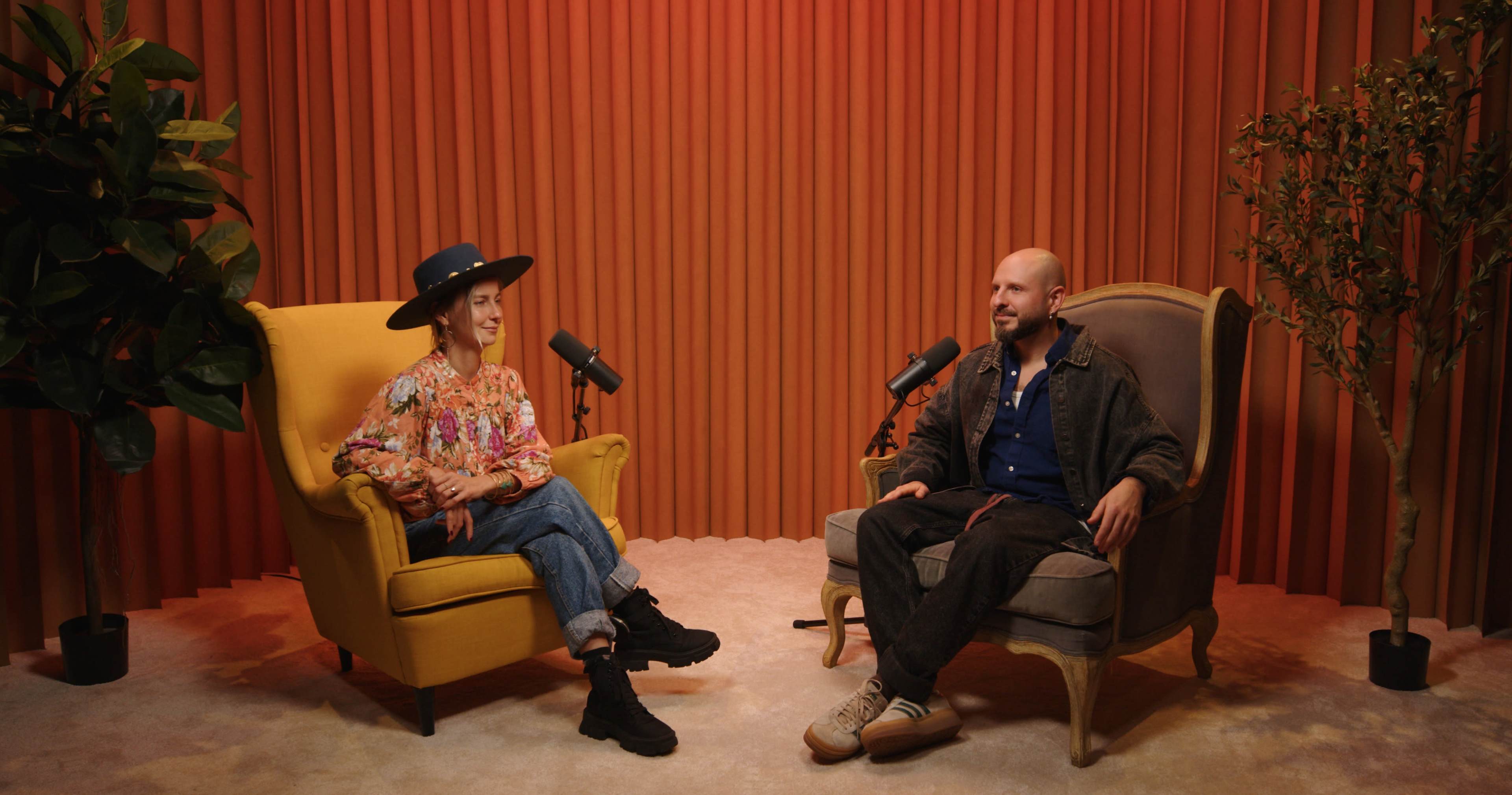 Two people are seated in armchairs in a studio setting, with microphones in front of them and a backdrop of vertical fabric panels.