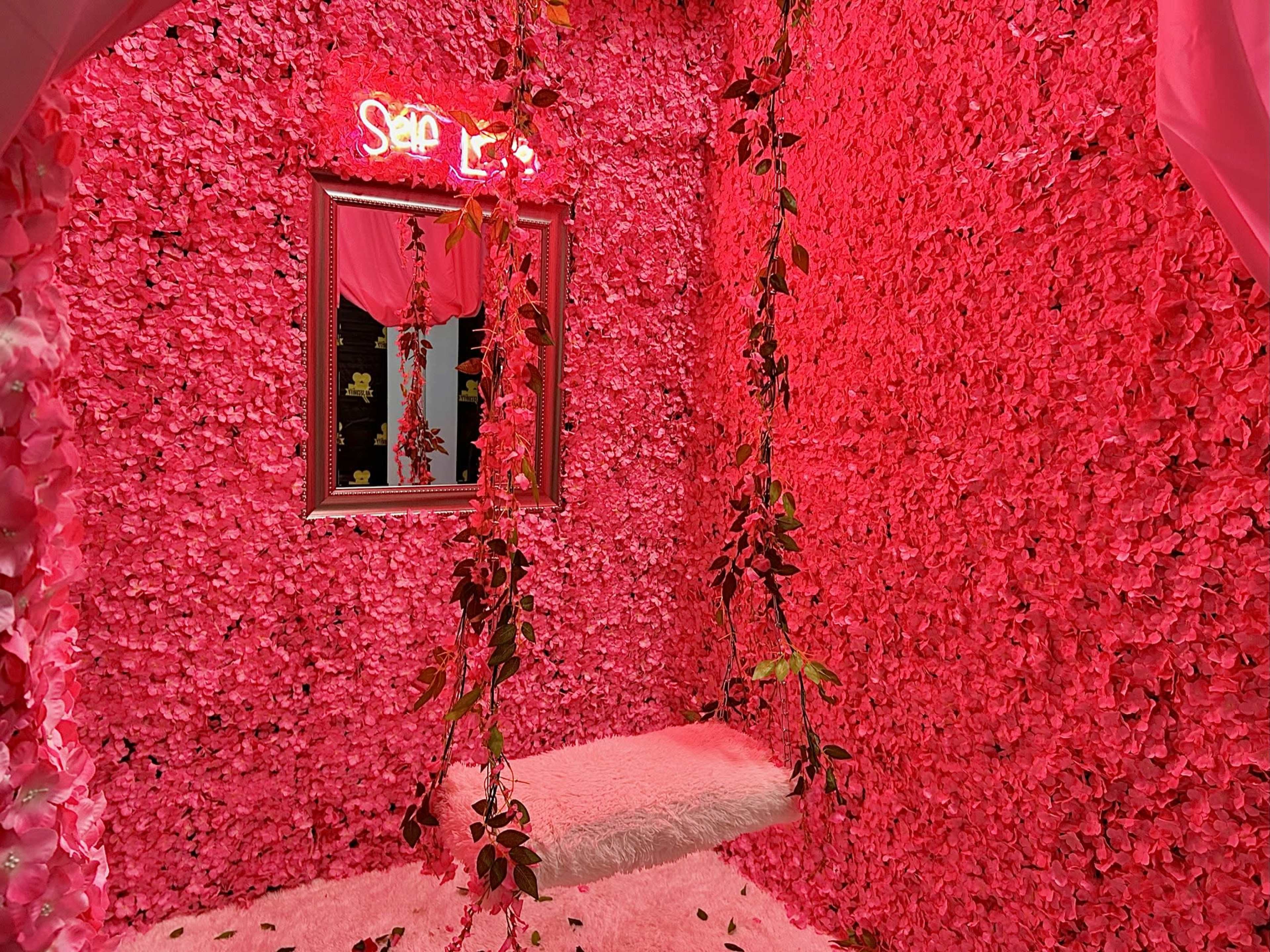 The room is adorned with pink floral walls, a swinging seat, and a neon sign that reads "Selfie."