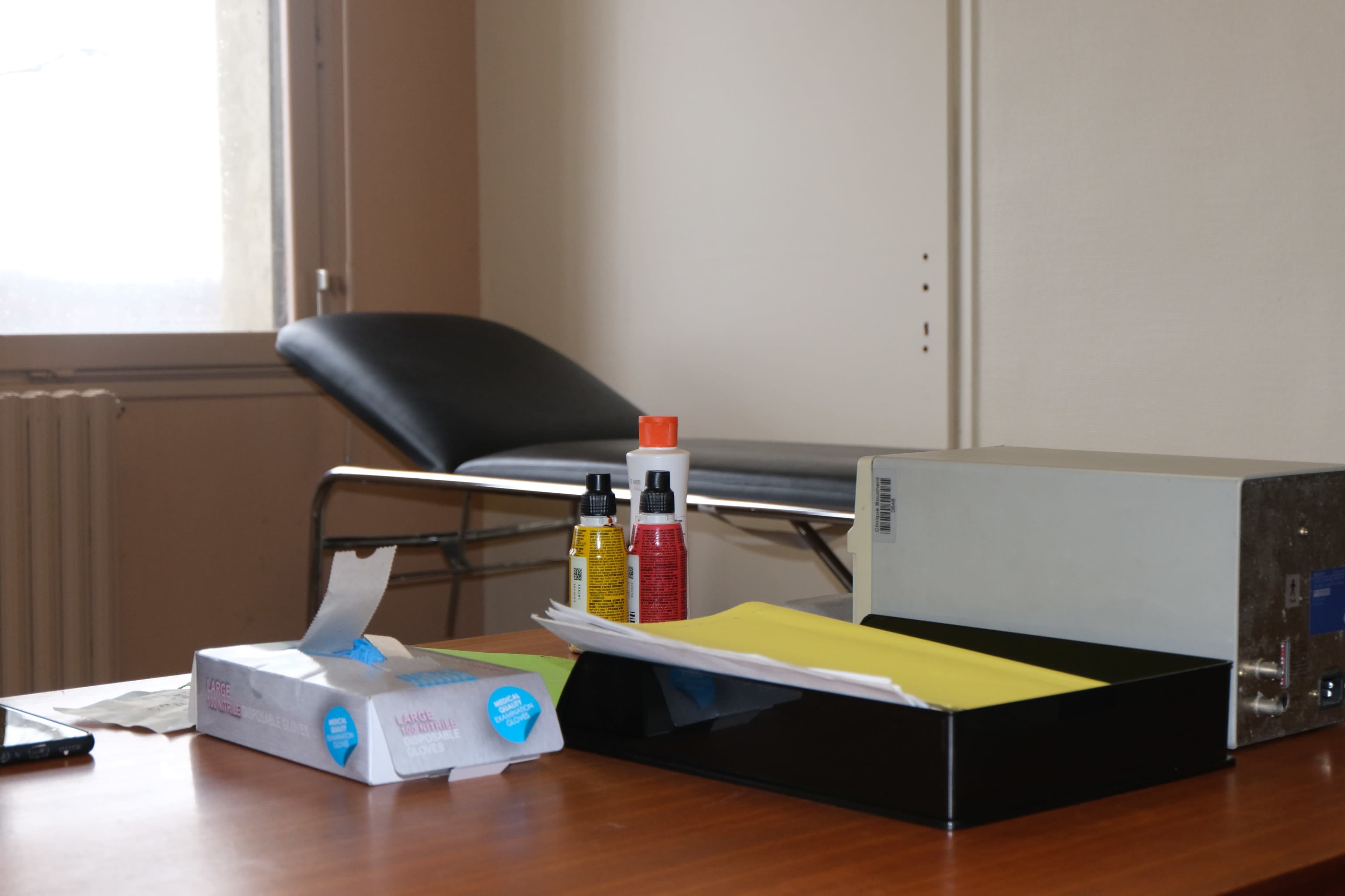 A small office space with a black examination table, a stack of yellow papers, and various bottles and items on a desk.