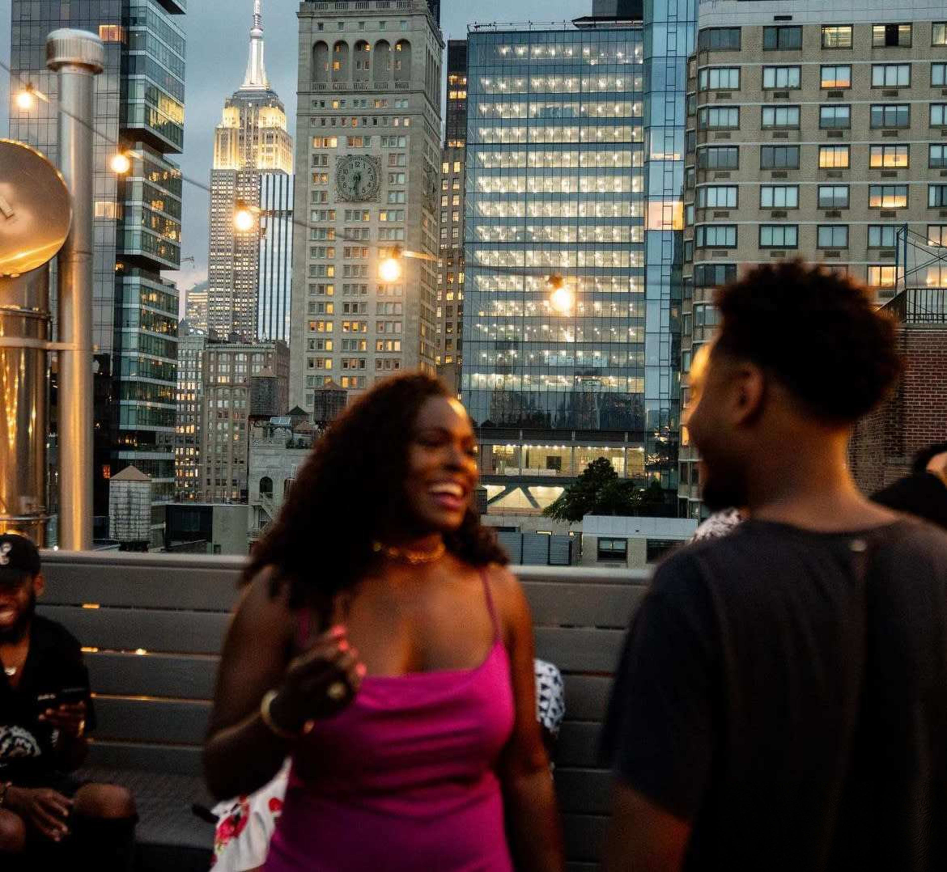 The image shows two people engaged in conversation on a rooftop bar with the Empire State Building visible in the background.