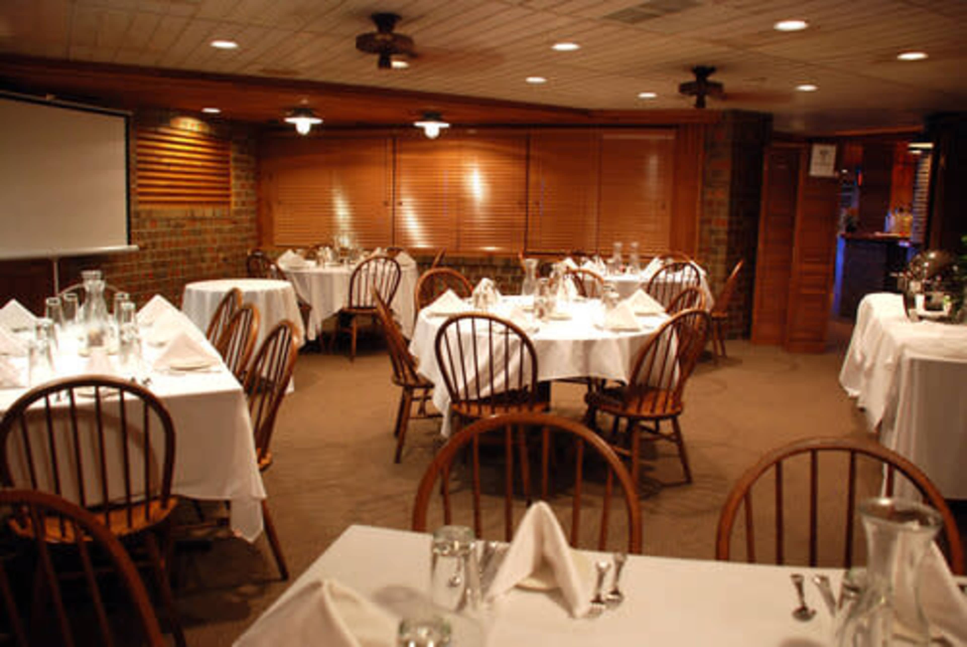 The image shows a dining room with several round tables set with white tablecloths and wooden chairs, illuminated by warm overhead lights.