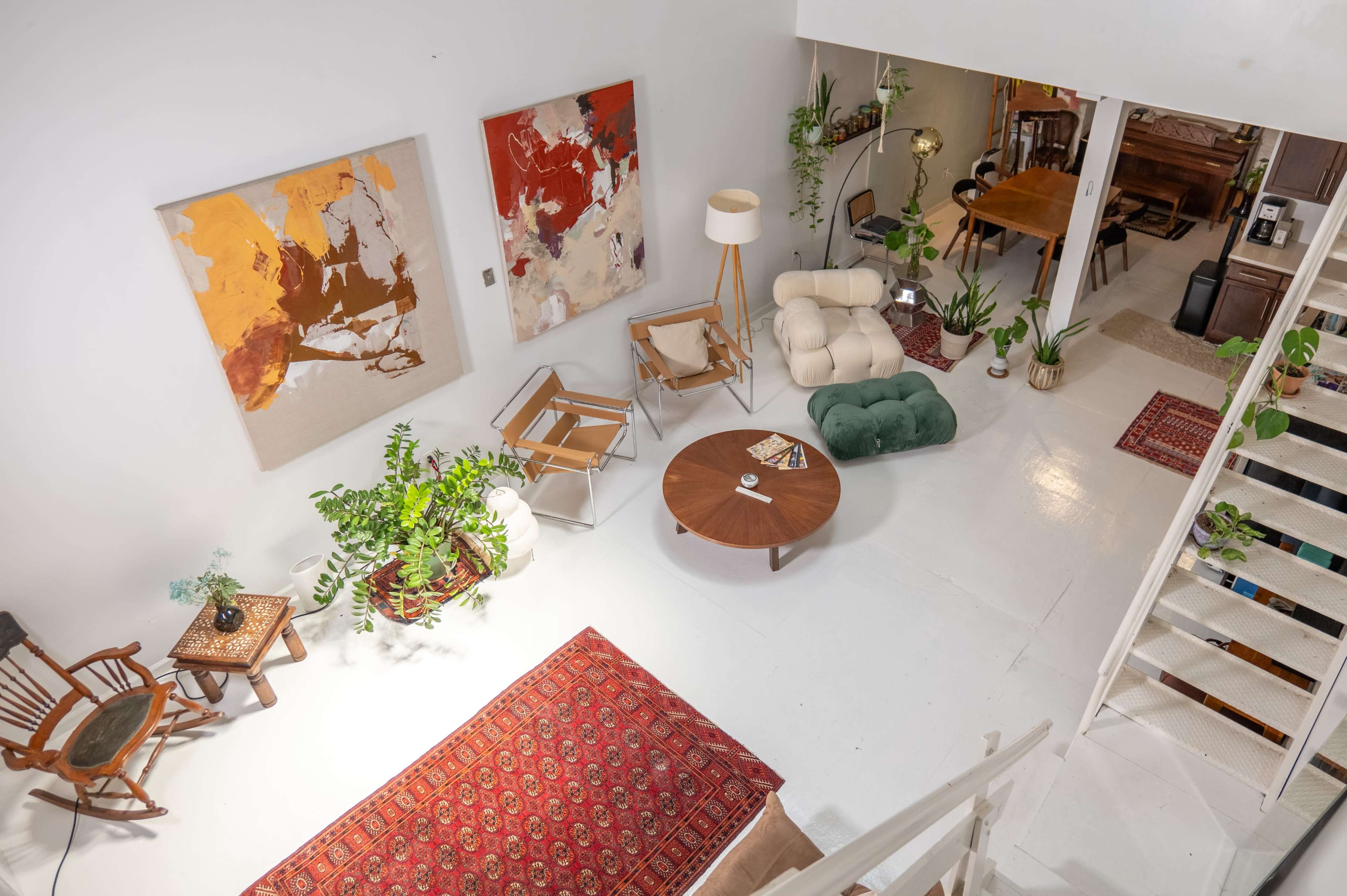 A bright, airy living space with a mix of modern furniture, plants, and artworks arranged around a circular coffee table on a white floor.