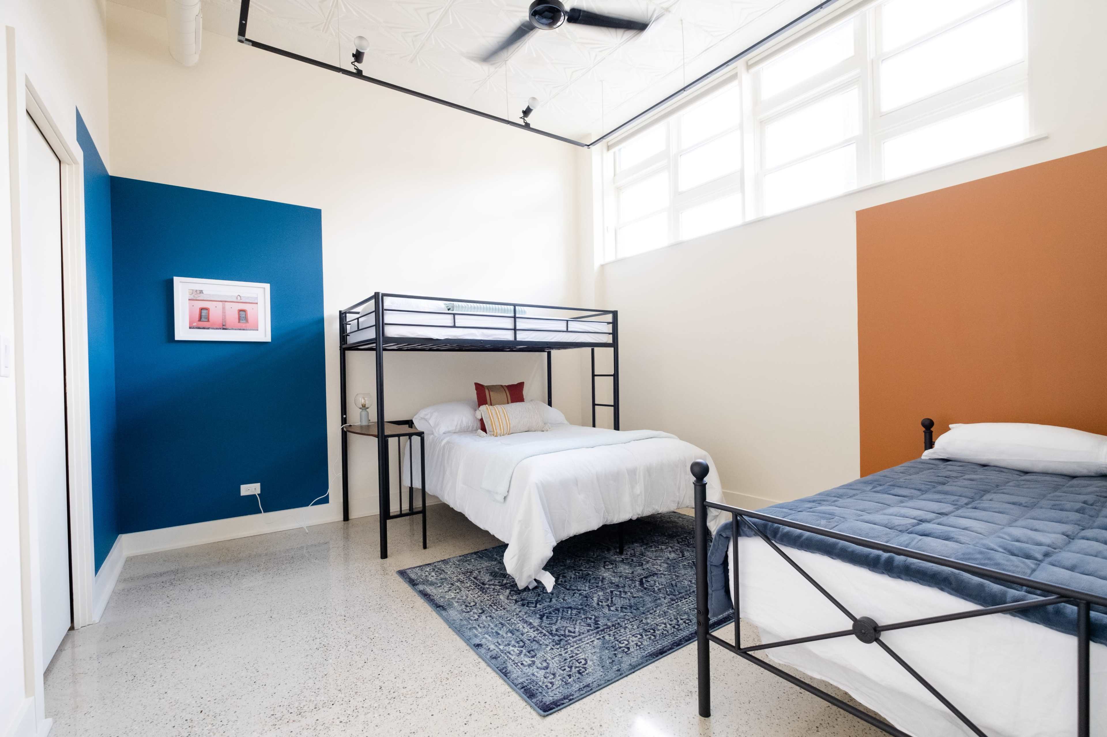 A modern bedroom with a bunk bed on the left, a single bed on the right, and a blue accent wall behind the bunk.