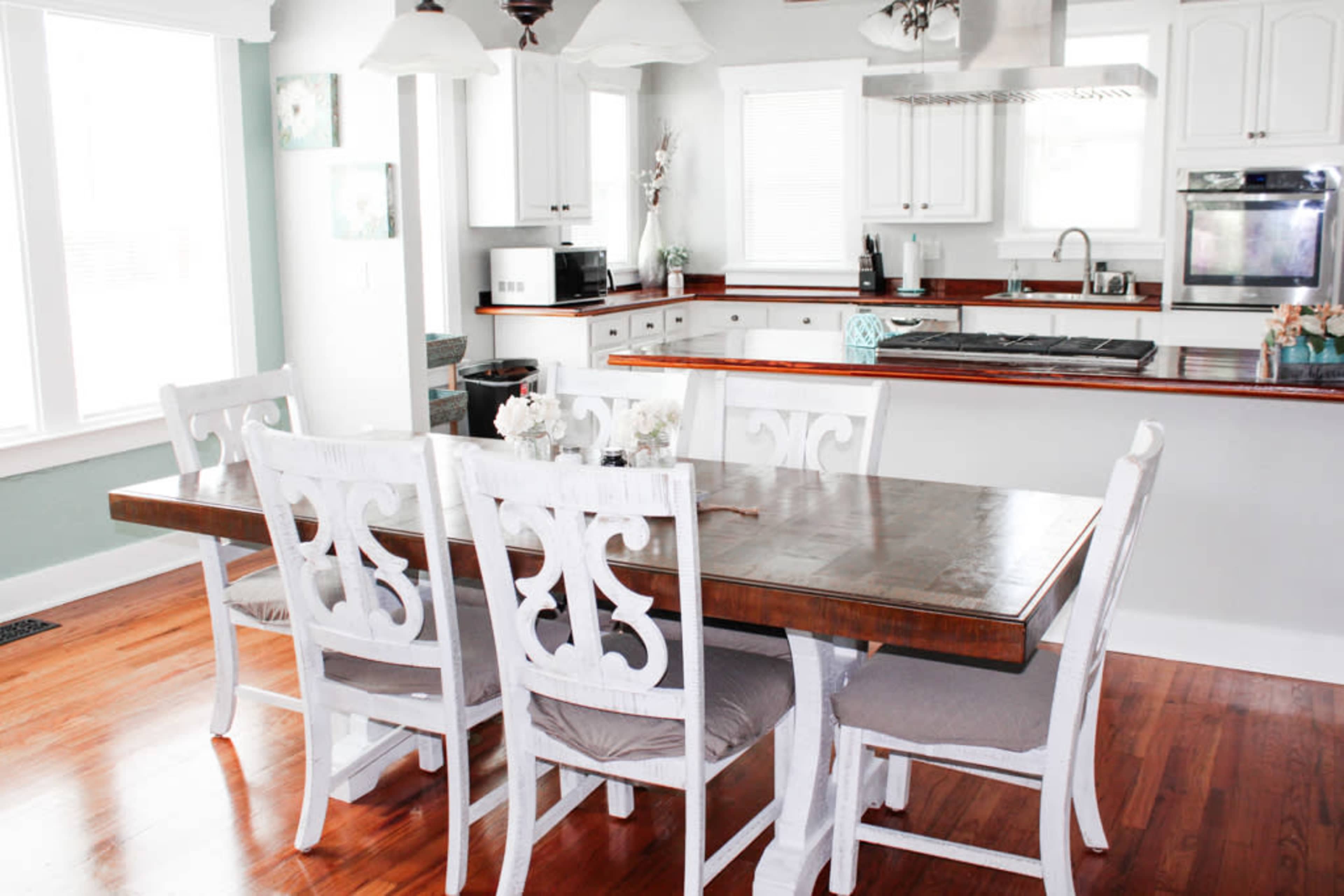 A dining table with six white chairs is set in a kitchen with wooden accents and light-colored cabinetry.