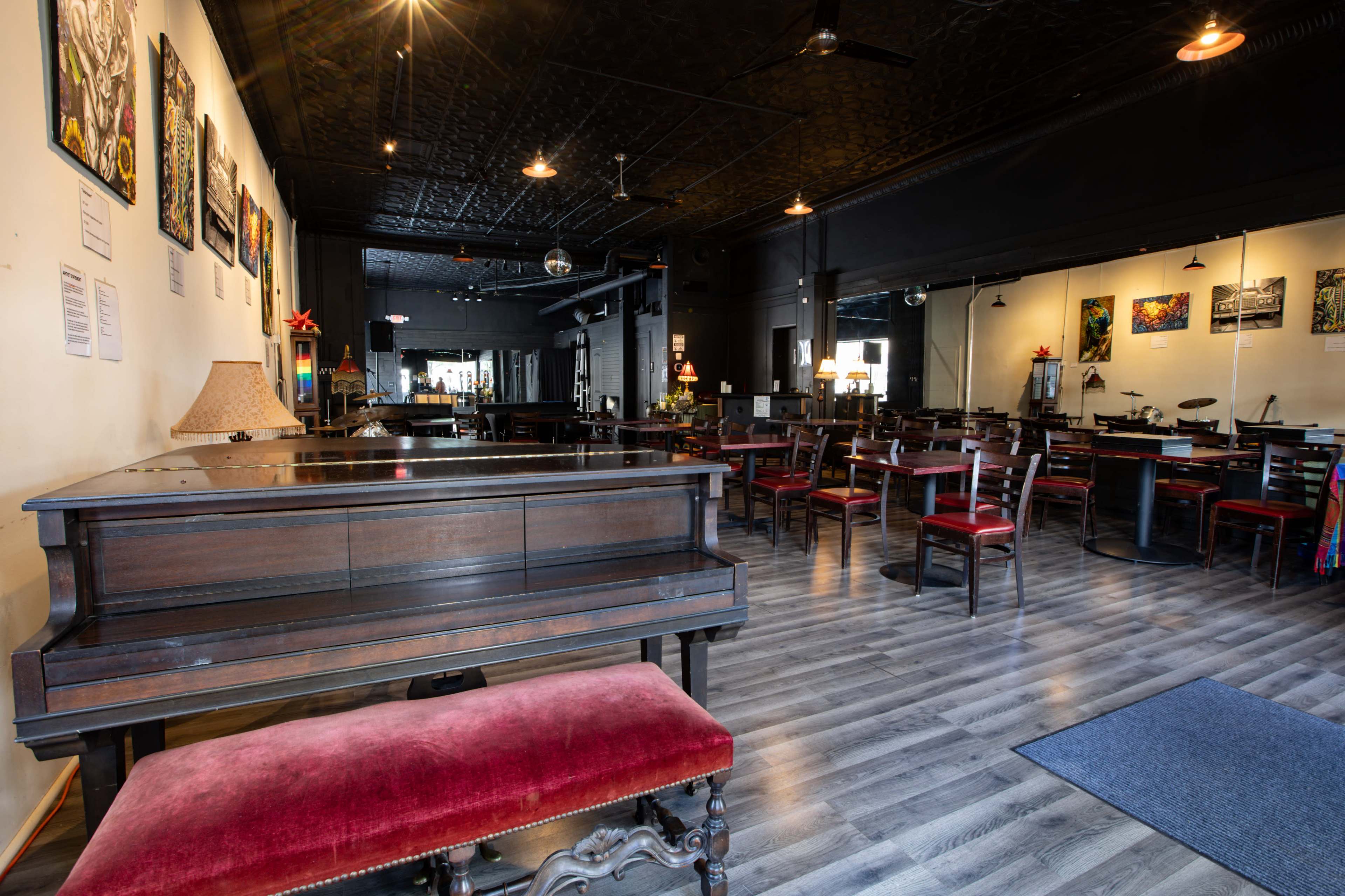 The image shows an interior of a restaurant or café featuring a wooden piano, red seating, and scattered tables with artwork displayed on the walls.
