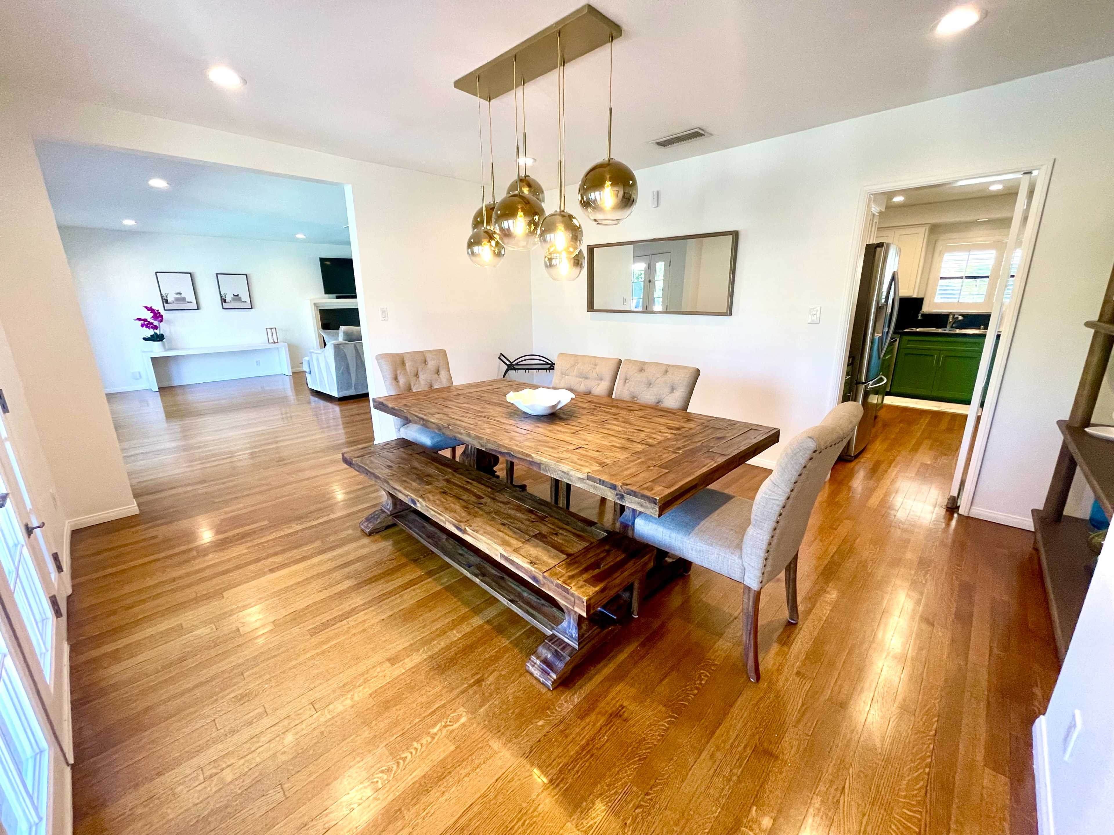 A wooden dining table with a long bench and upholstered chairs is situated in a bright, open dining area with pendant lighting above.
