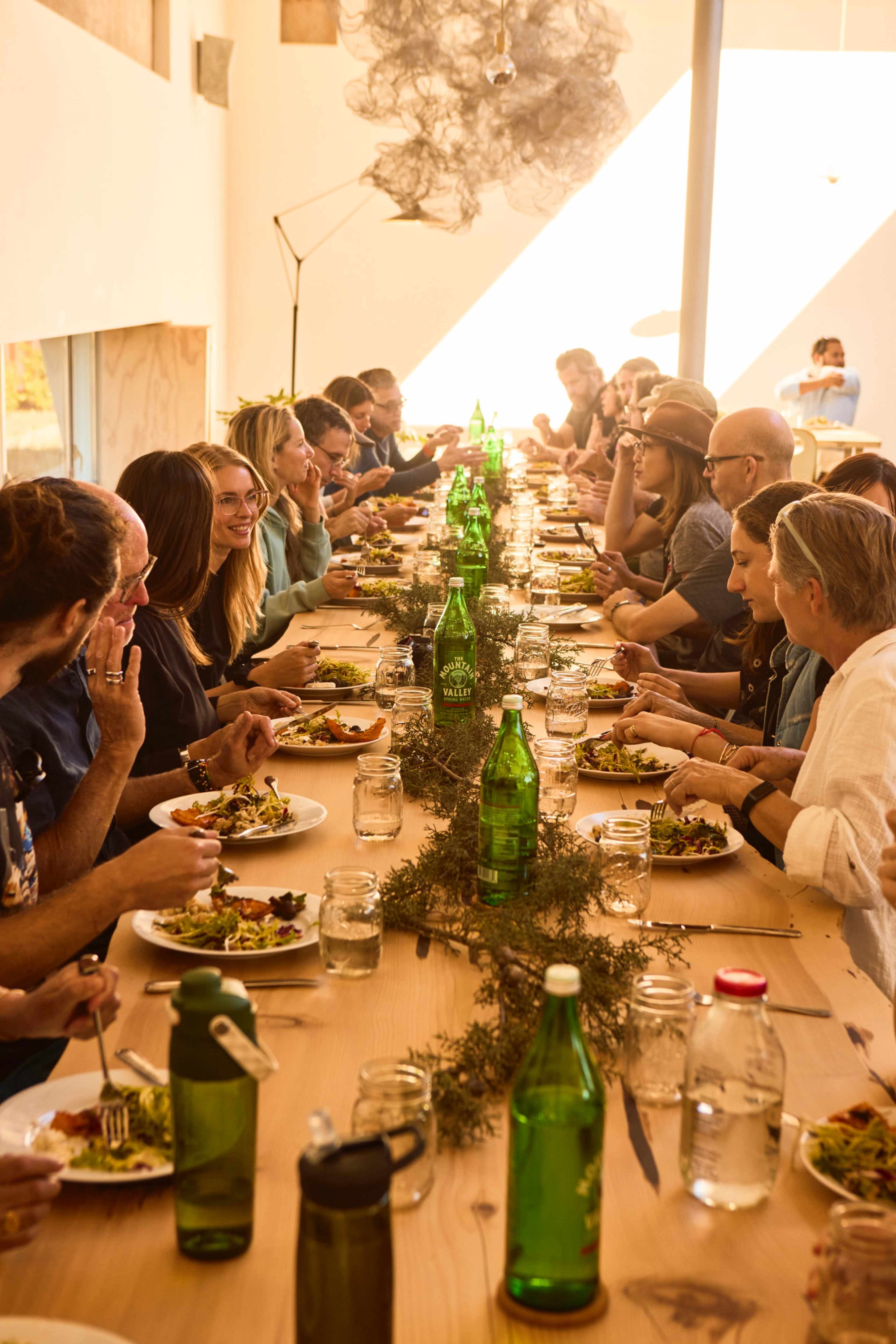 A large group of people sits at a long table, sharing a meal with plates of food and green bottles of water in a bright, spacious room.