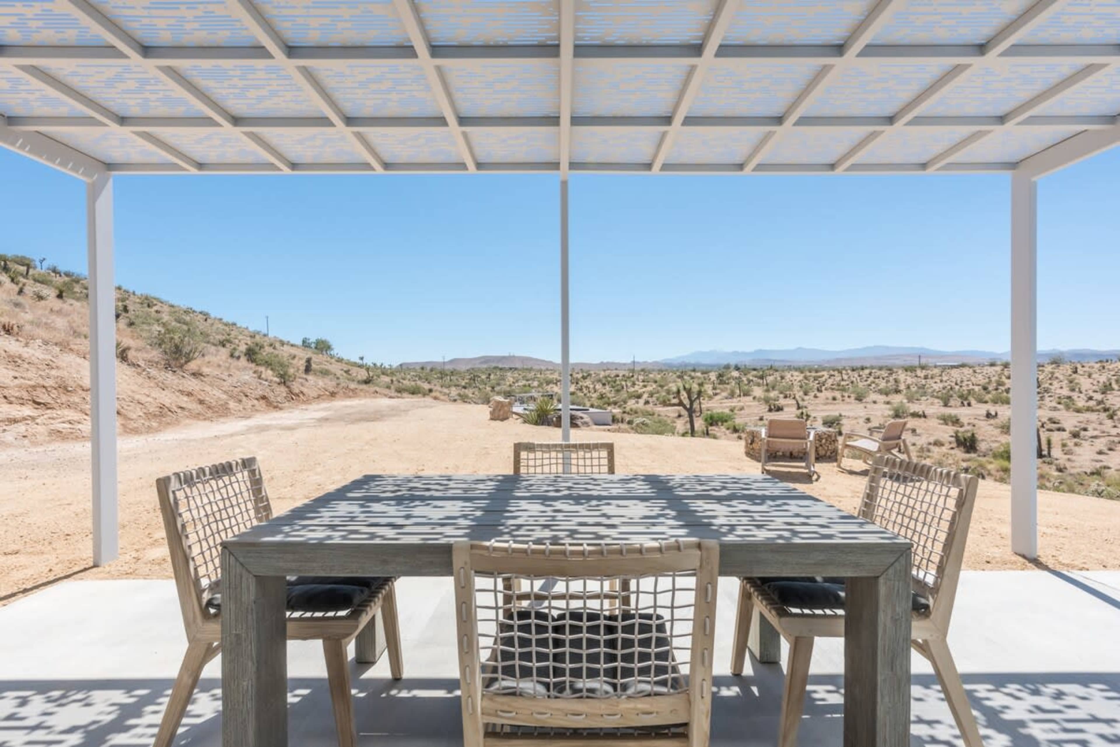 A large outdoor dining table with chairs is set under a white pergola, overlooking a desert landscape with sparse vegetation and distant mountains.