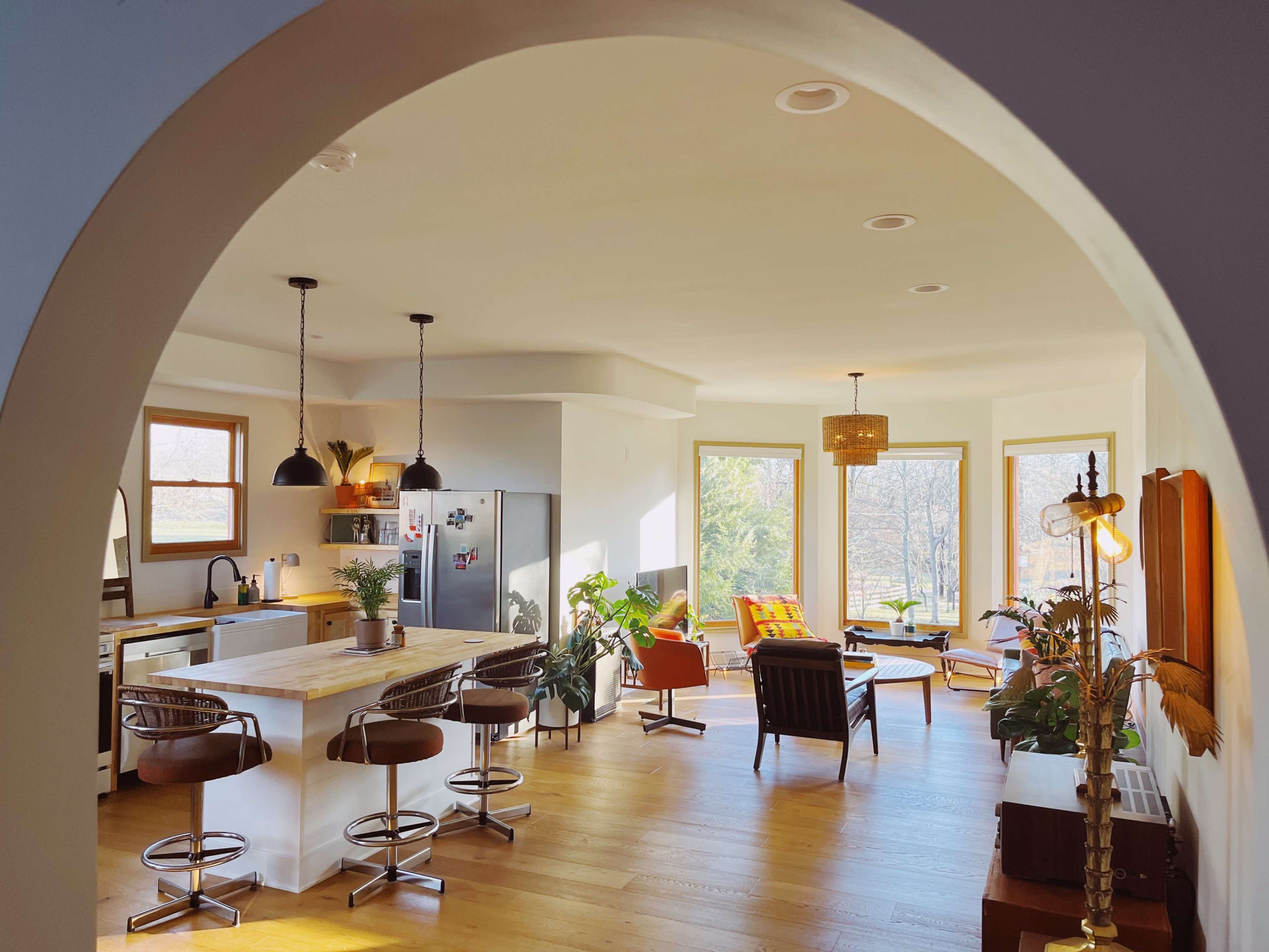 A bright, open-concept living area and kitchen with large windows, modern furniture, and plants.