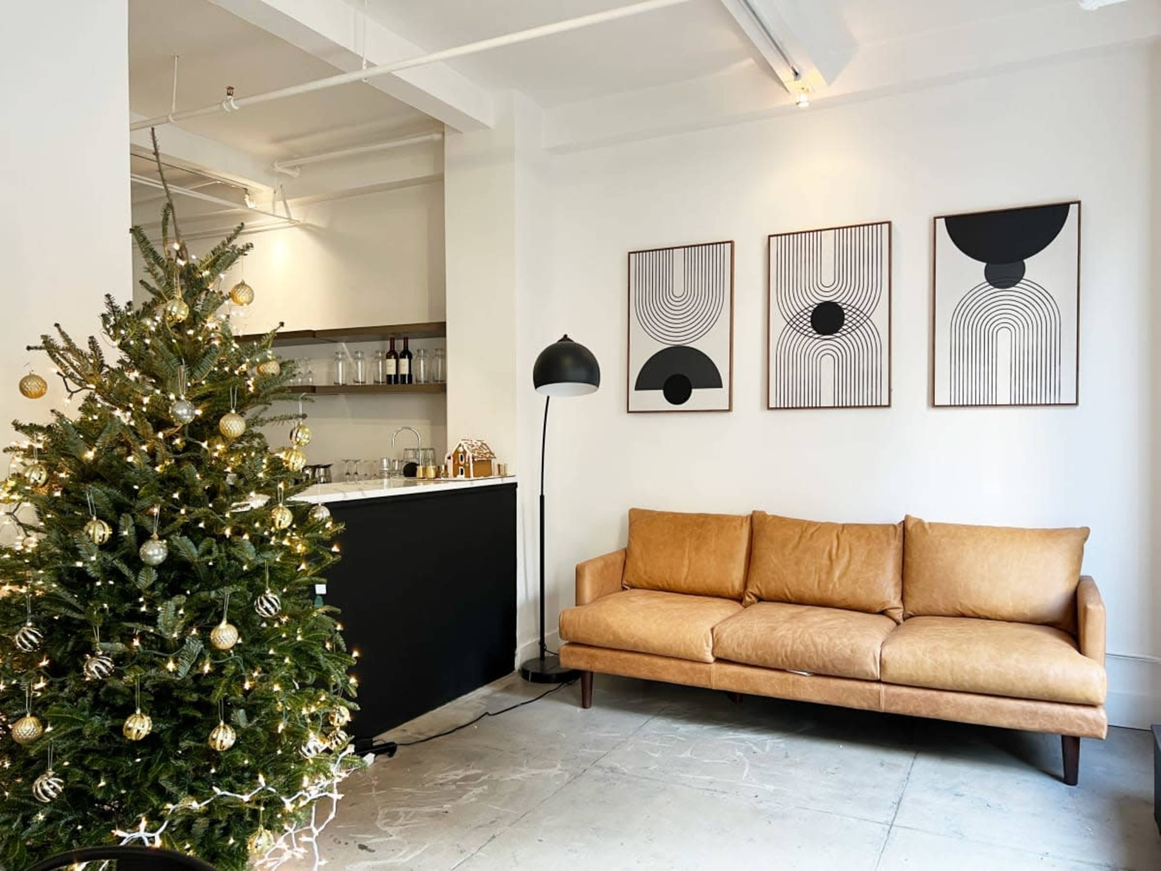 A decorated Christmas tree stands next to a brown leather sofa and three abstract framed artworks on a white wall in a modern living space.
