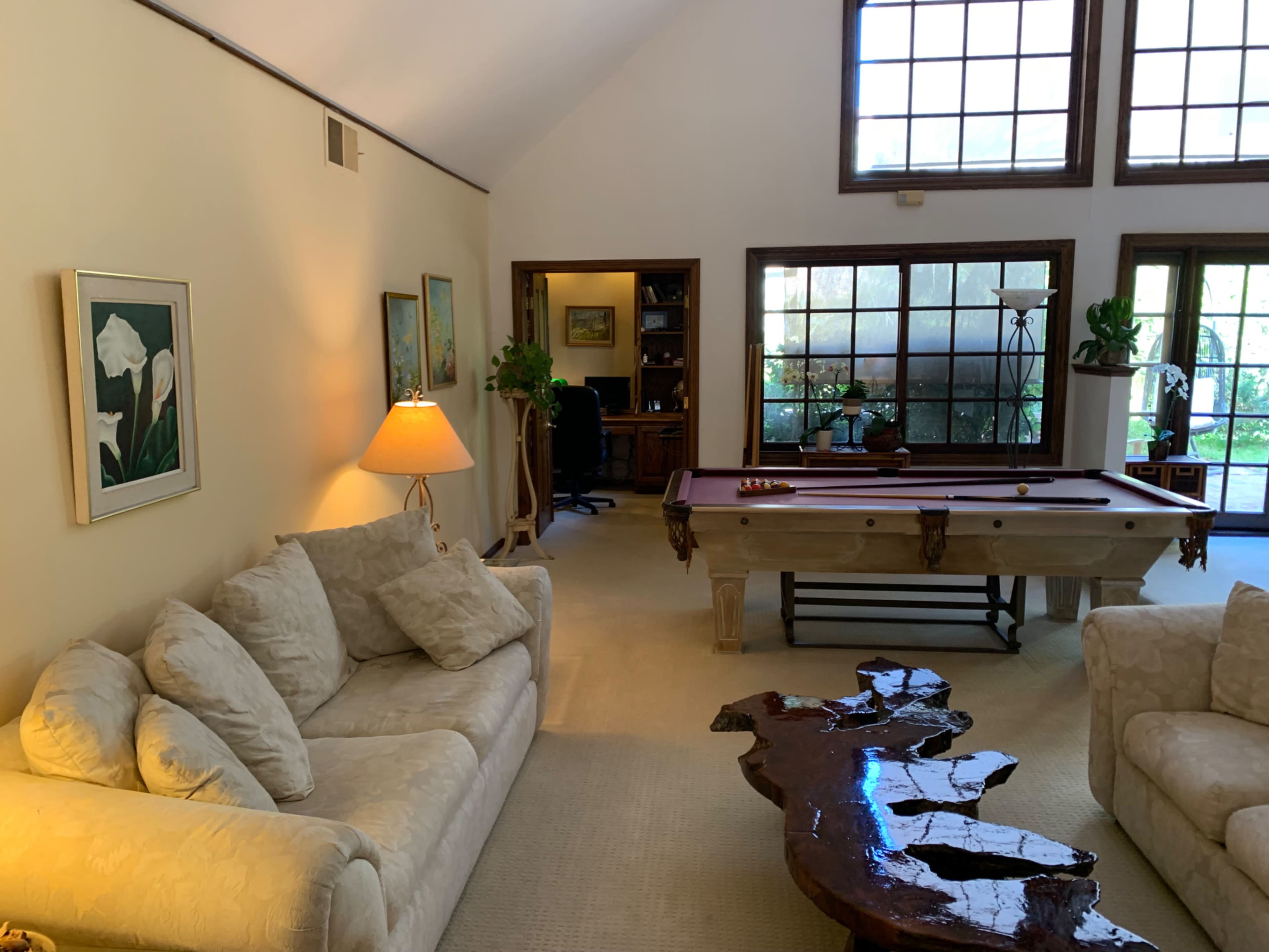 The image shows a spacious room with a beige sofa, a wooden pool table, and large windows providing natural light.