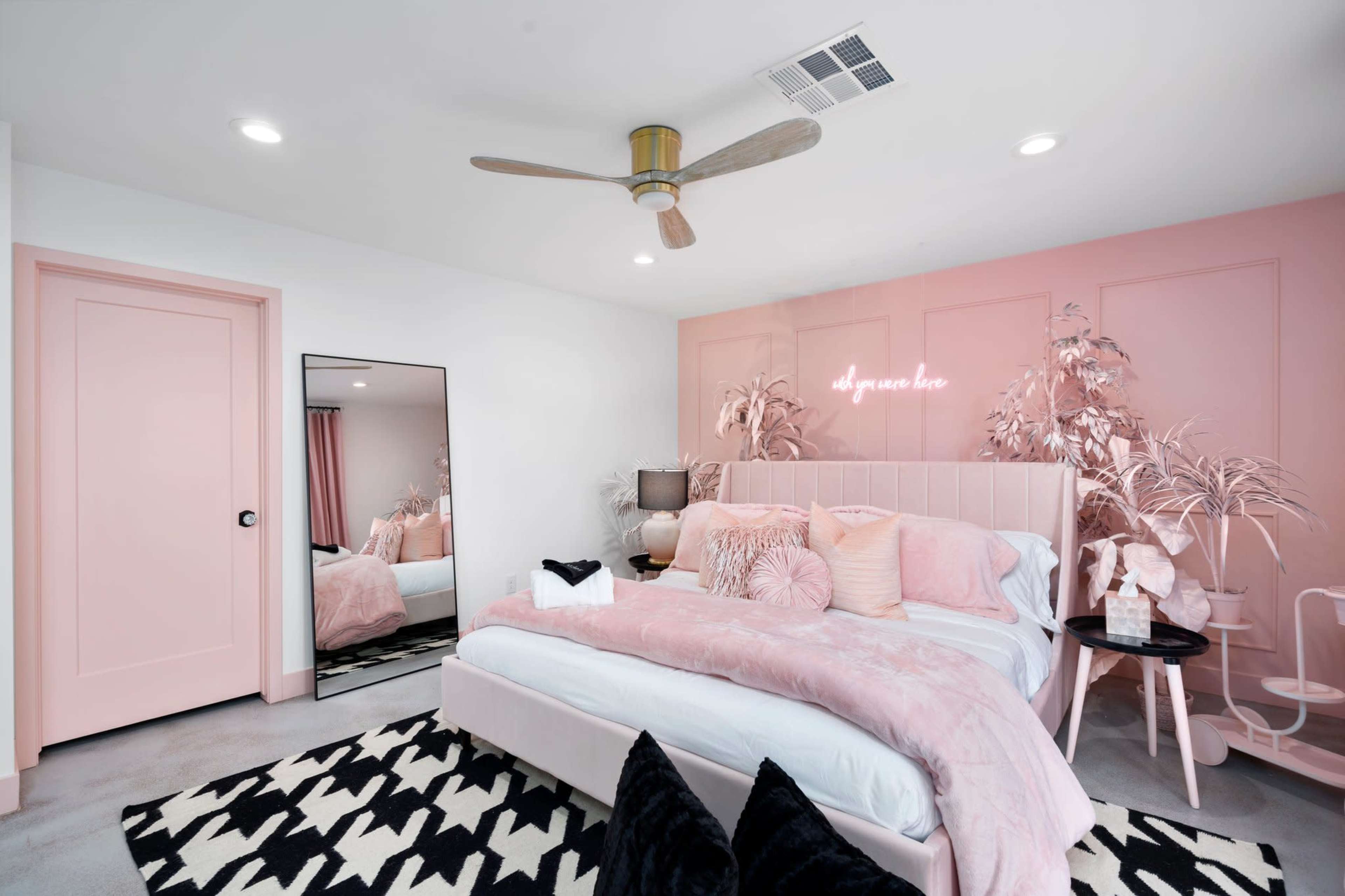 The image shows a modern bedroom with a pink and white color scheme, featuring a large bed, a mirror, a ceiling fan, and a decorative rug.