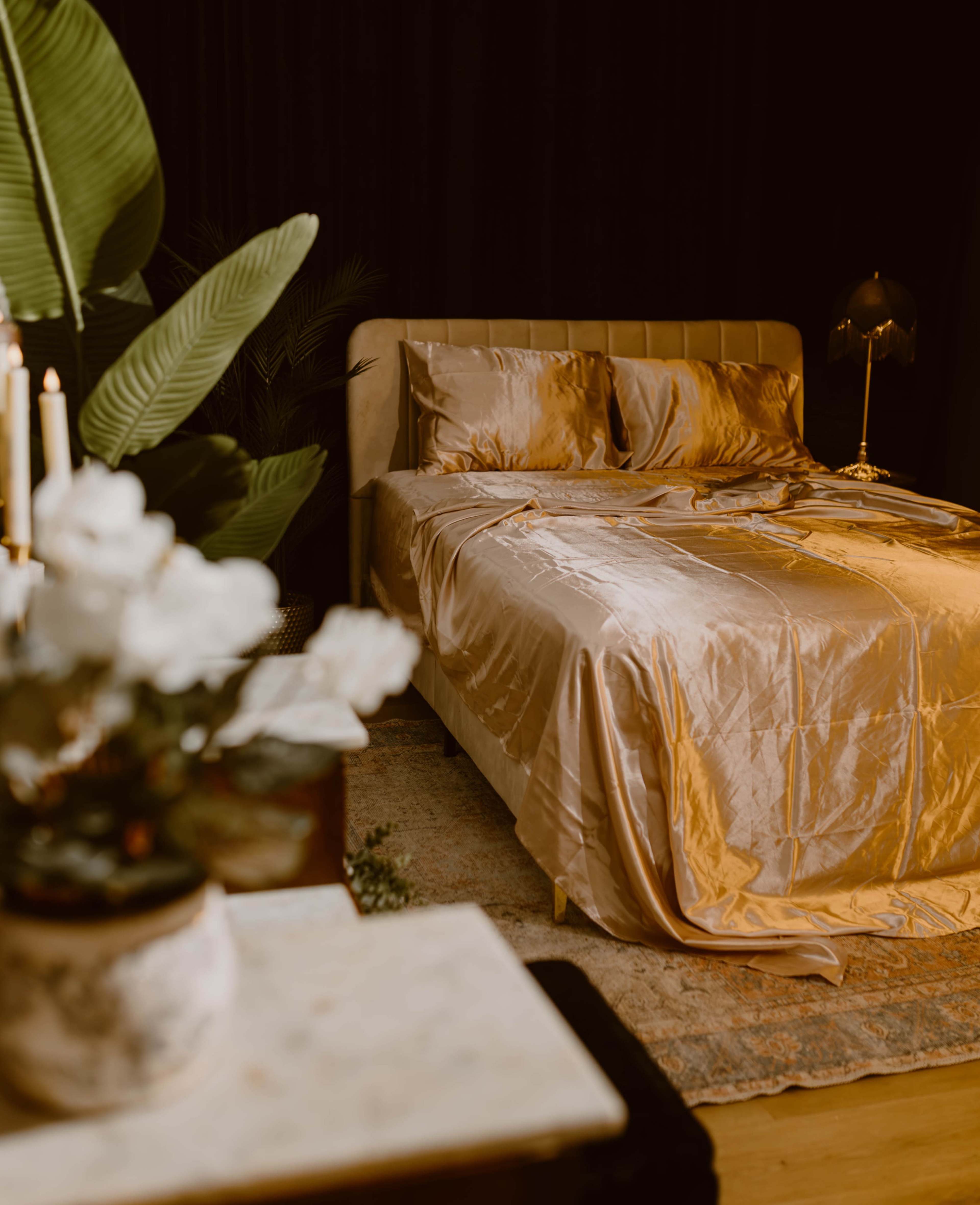 The image shows a neatly made bed with a silky golden comforter, surrounded by dark curtains and a lamp on the nightstand, complemented by a decorative plant and flowers in the foreground.