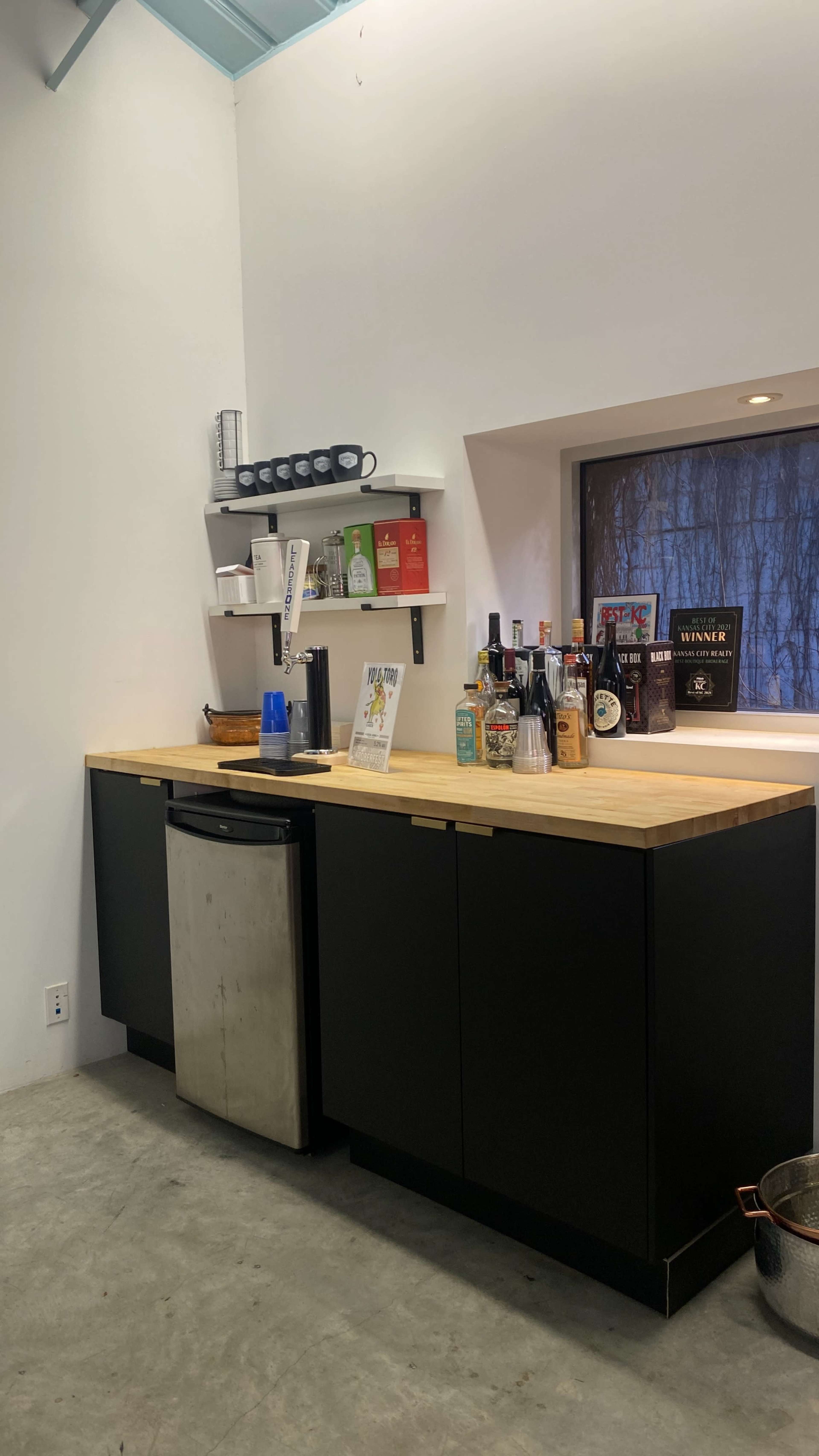 A minimalist kitchen bar with a wooden countertop, black cabinets, and a variety of bottles displayed on shelves and the counter.