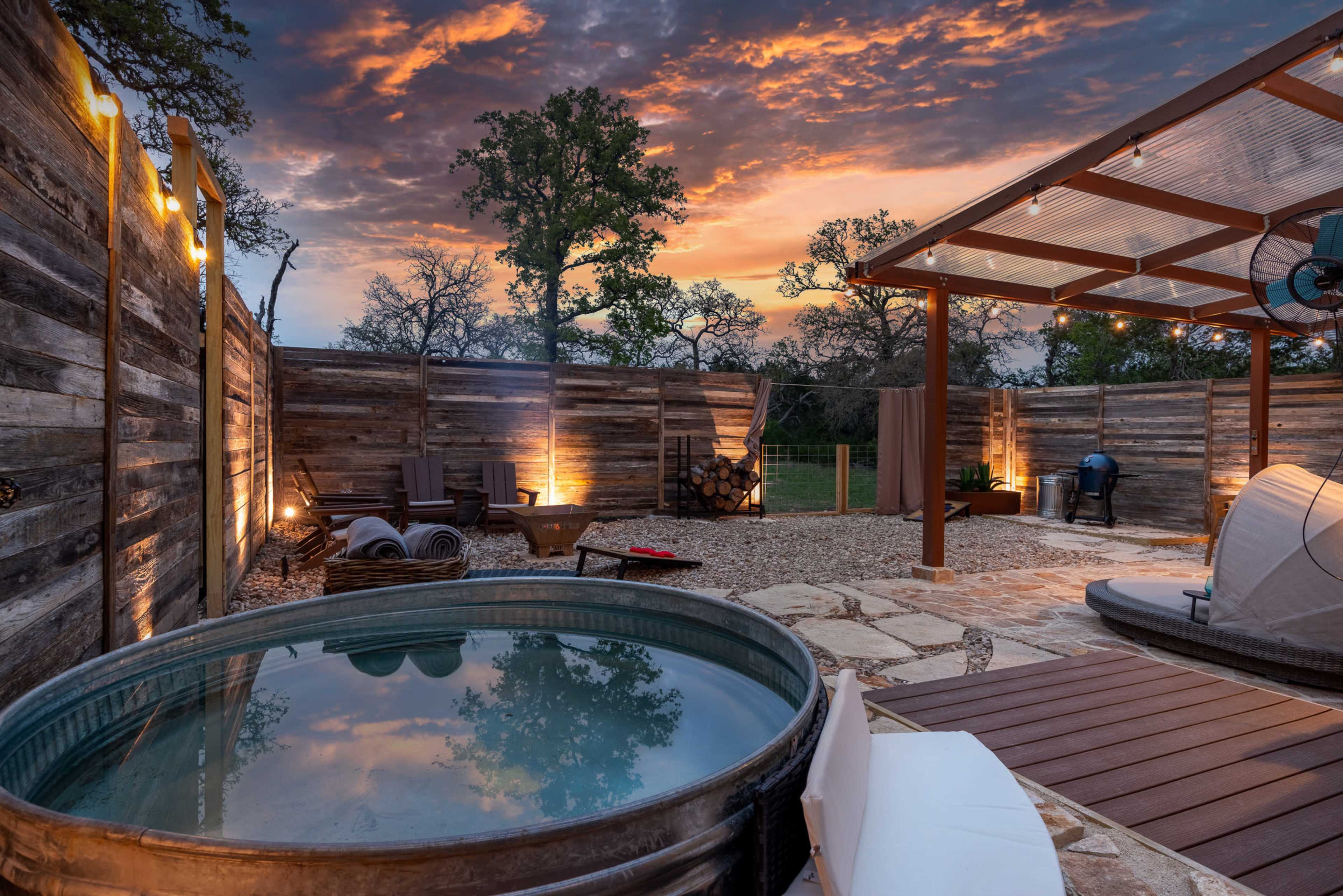 A wooden hot tub sits on a stone patio surrounded by rustic fencing, with a scenic sunset sky visible above.