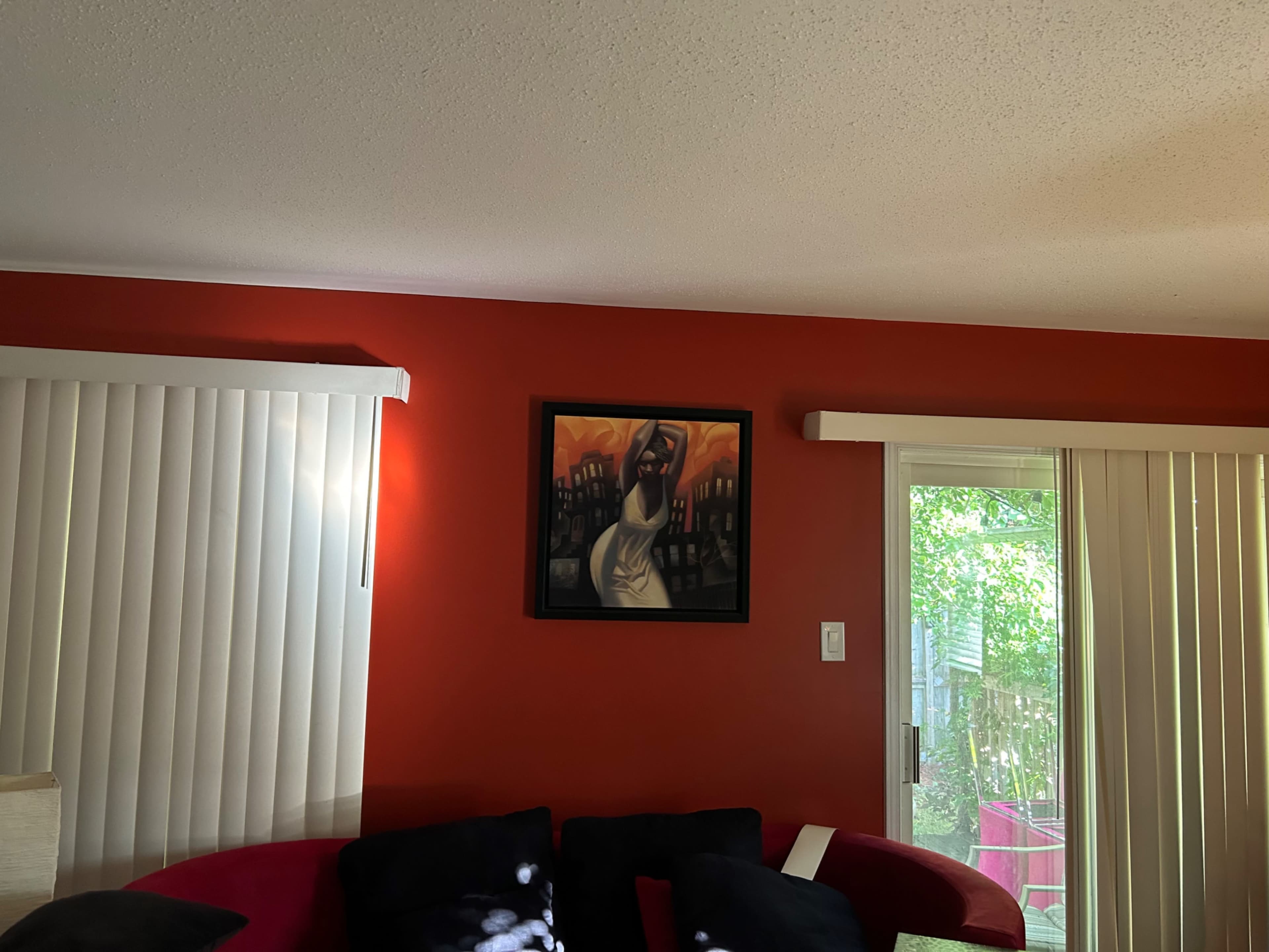 A framed painting of a woman is displayed on a red wall next to a window with vertical blinds.
