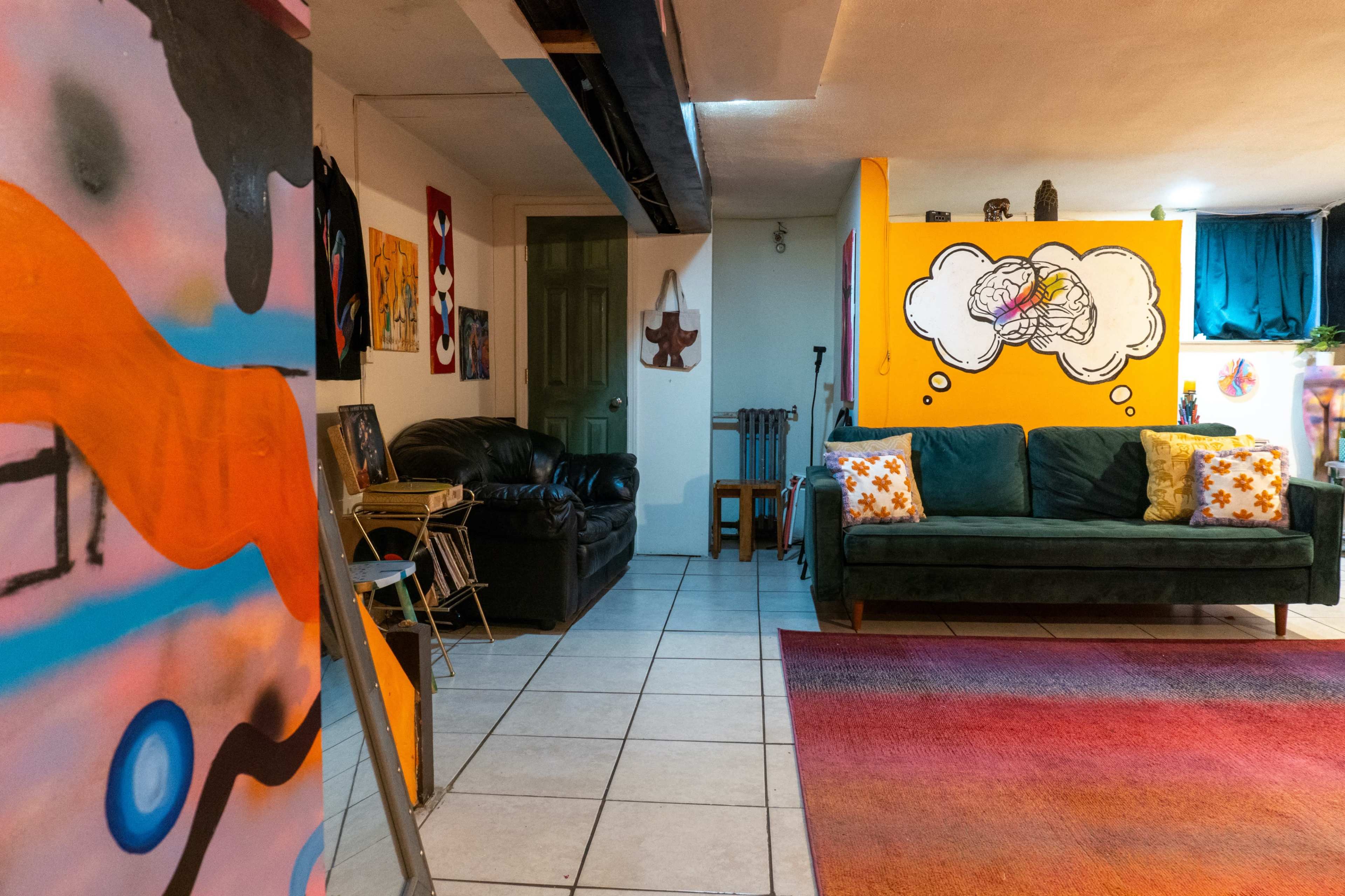 The image shows a colorful basement lounge area featuring a dark sofa, a green couch with decorative pillows, an orange wall with a wall mural, and various artworks hanging on the walls.