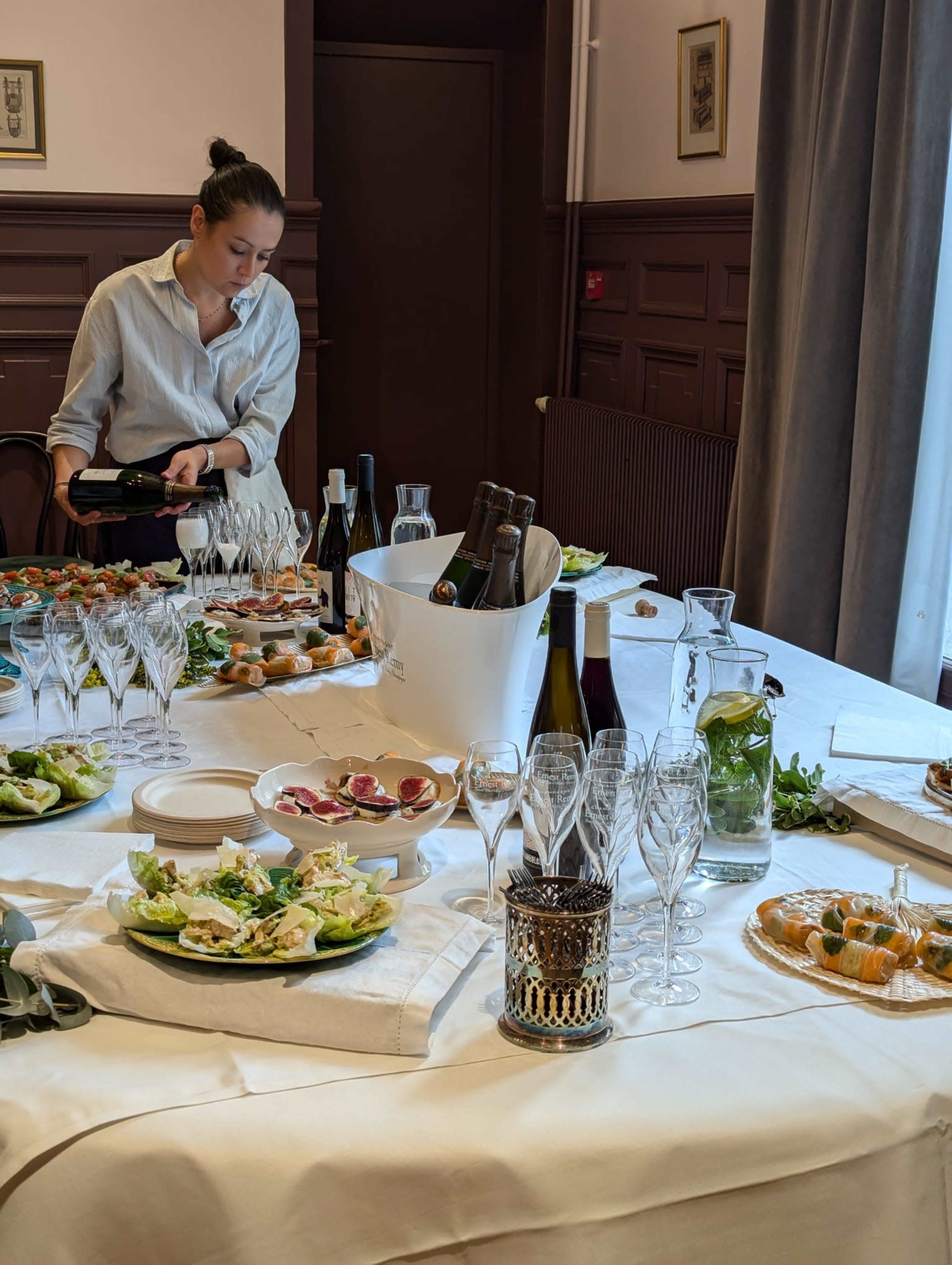 A woman prepares a table set with various dishes, drinks, and glasses for an event.