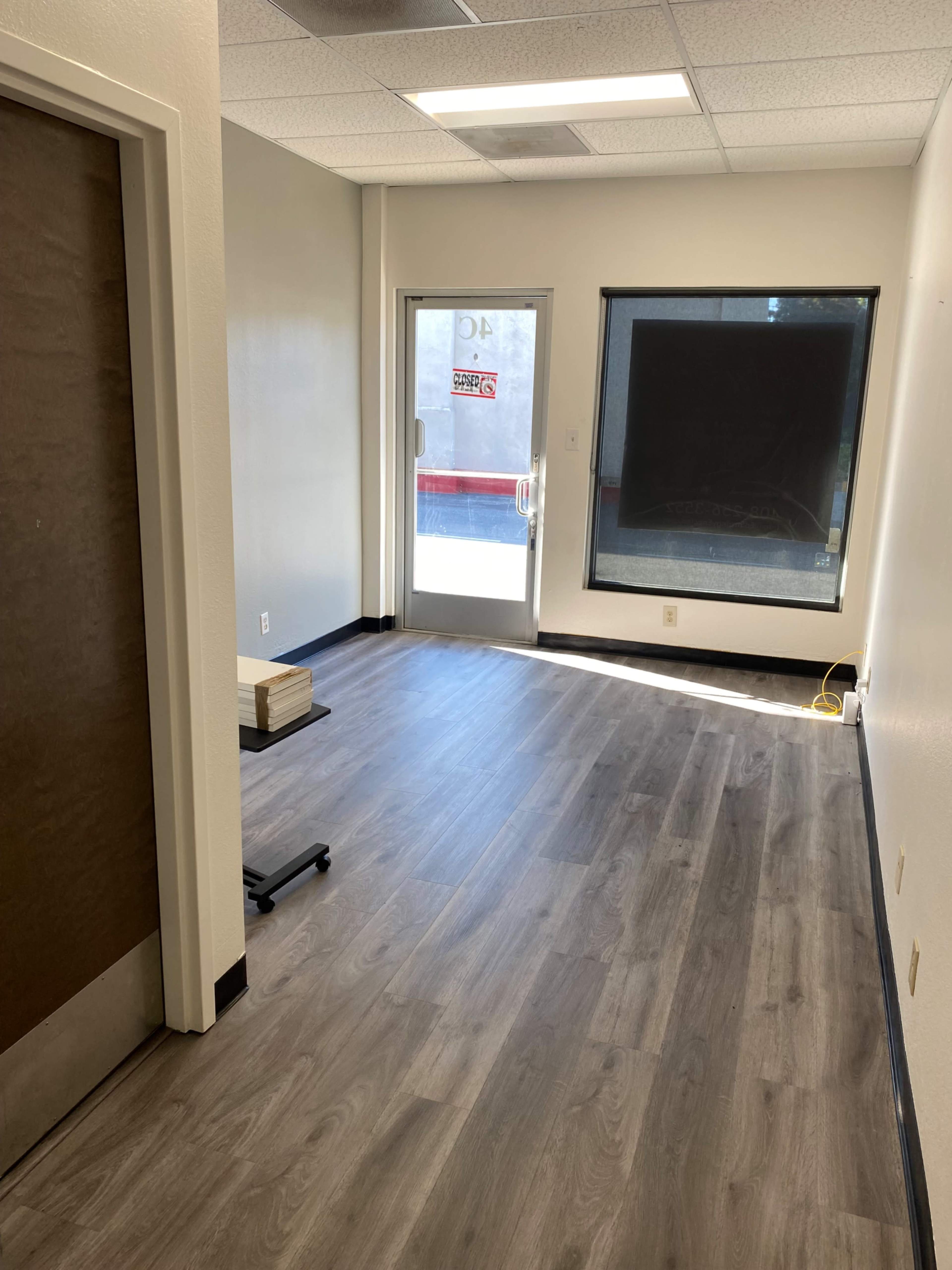 An empty room with light-colored walls, a door leading outside, and large windows, featuring gray laminate flooring.