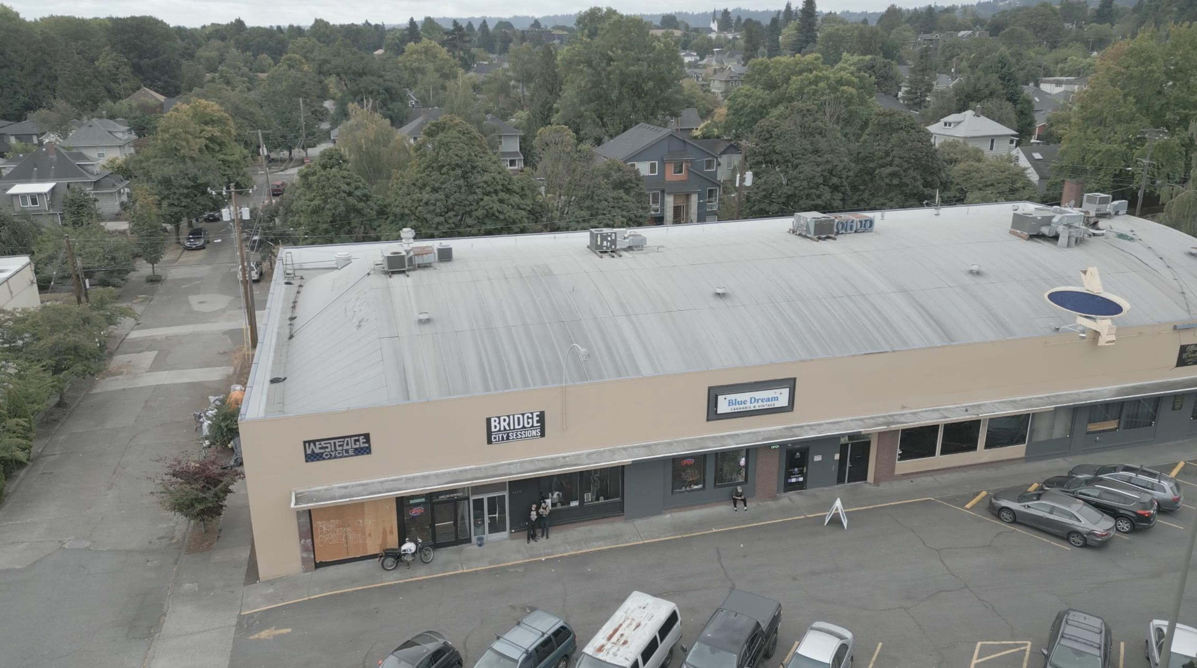 The image shows a large commercial building with multiple storefronts, surrounded by parked cars and residential houses in the background.