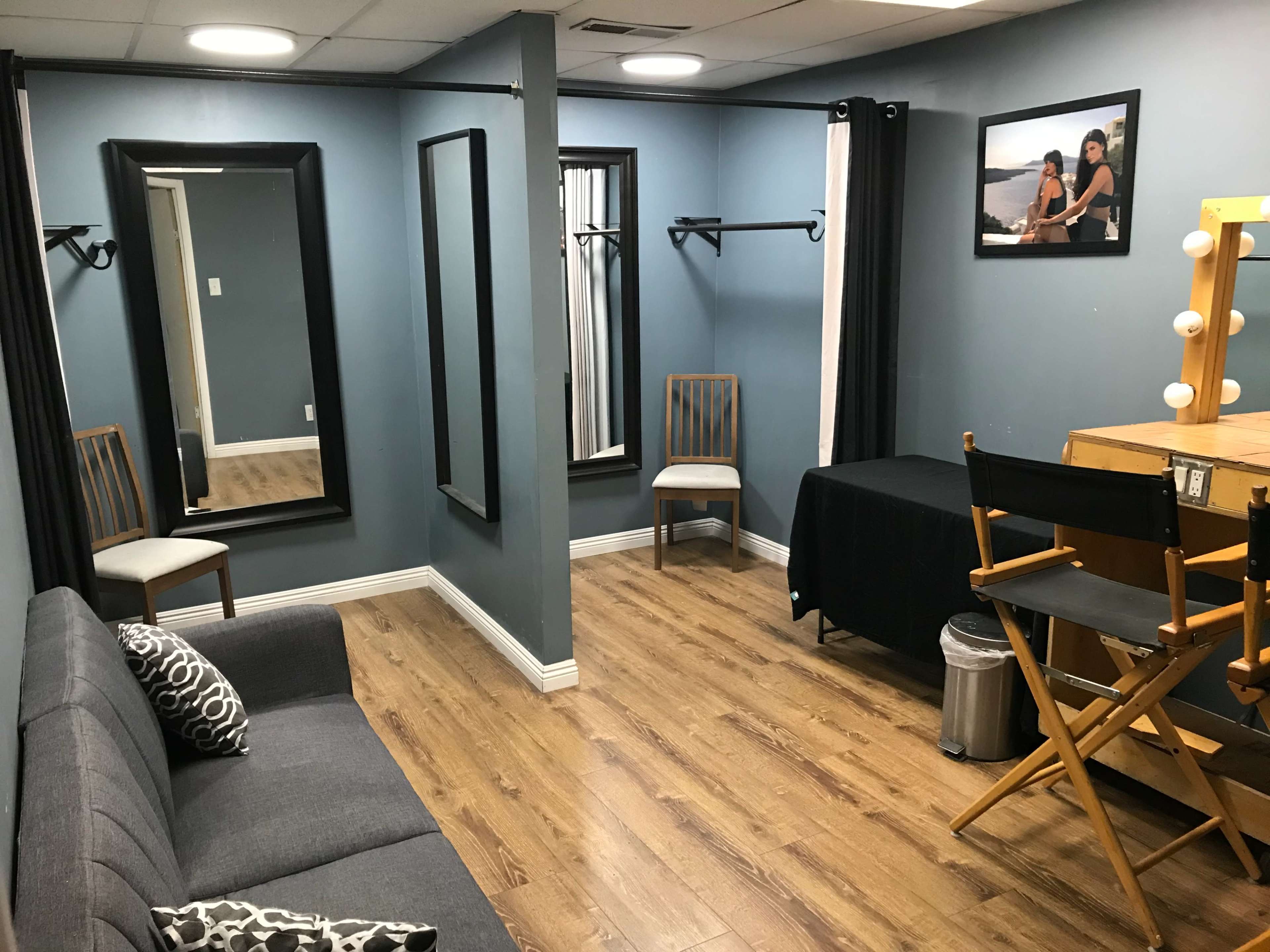 A dressing room with a gray sofa, a makeup station featuring a mirror and lights, two chairs, and three mirrors against walls painted blue.