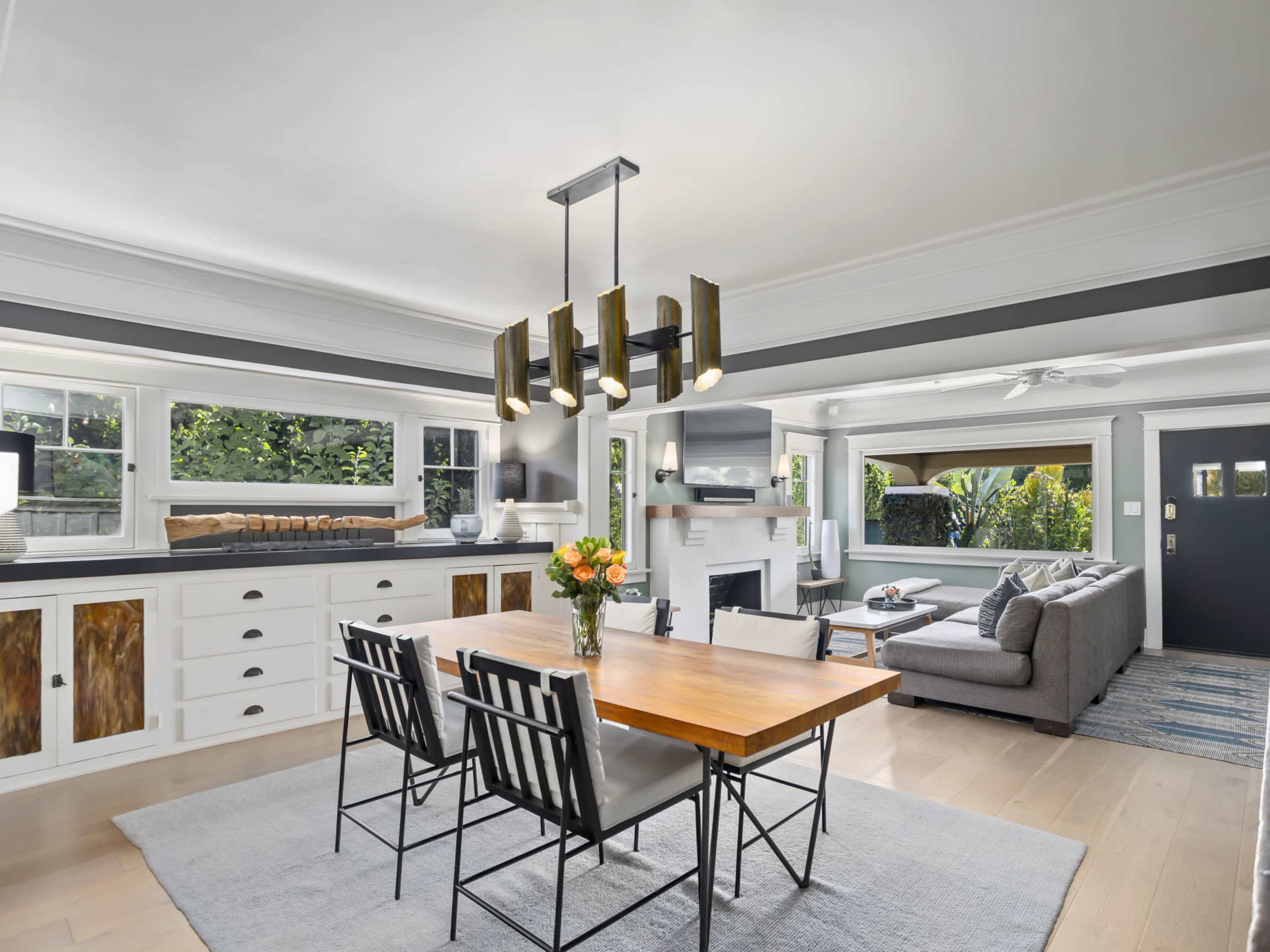 The image shows a modern dining area with a wooden table, metal chairs, a chandelier, and a view into a living room featuring a sofa and large windows.