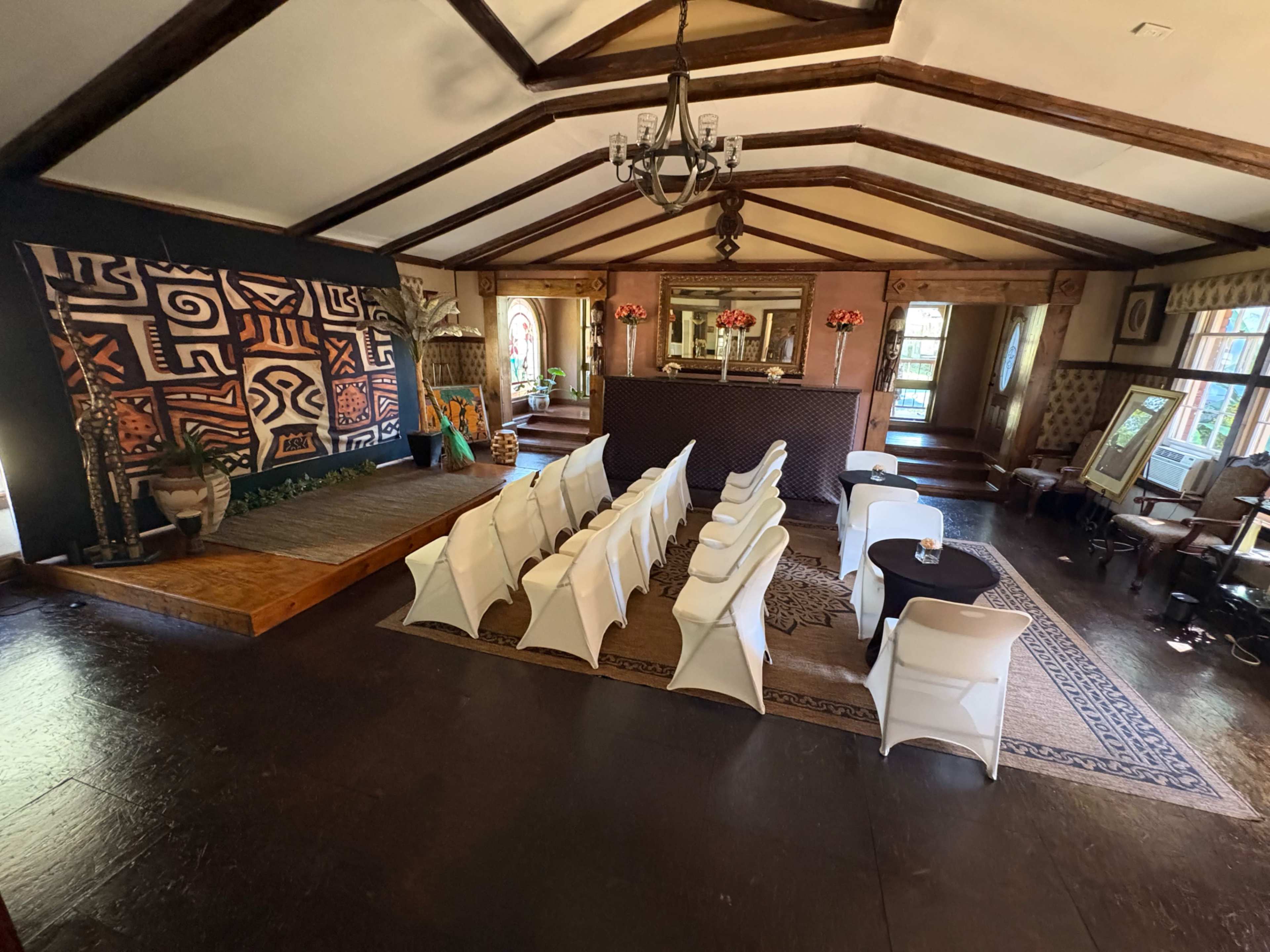 The image shows a spacious event room with a wooden beam ceiling, rows of white chairs set up for a gathering, and a large decorative mural on one wall.