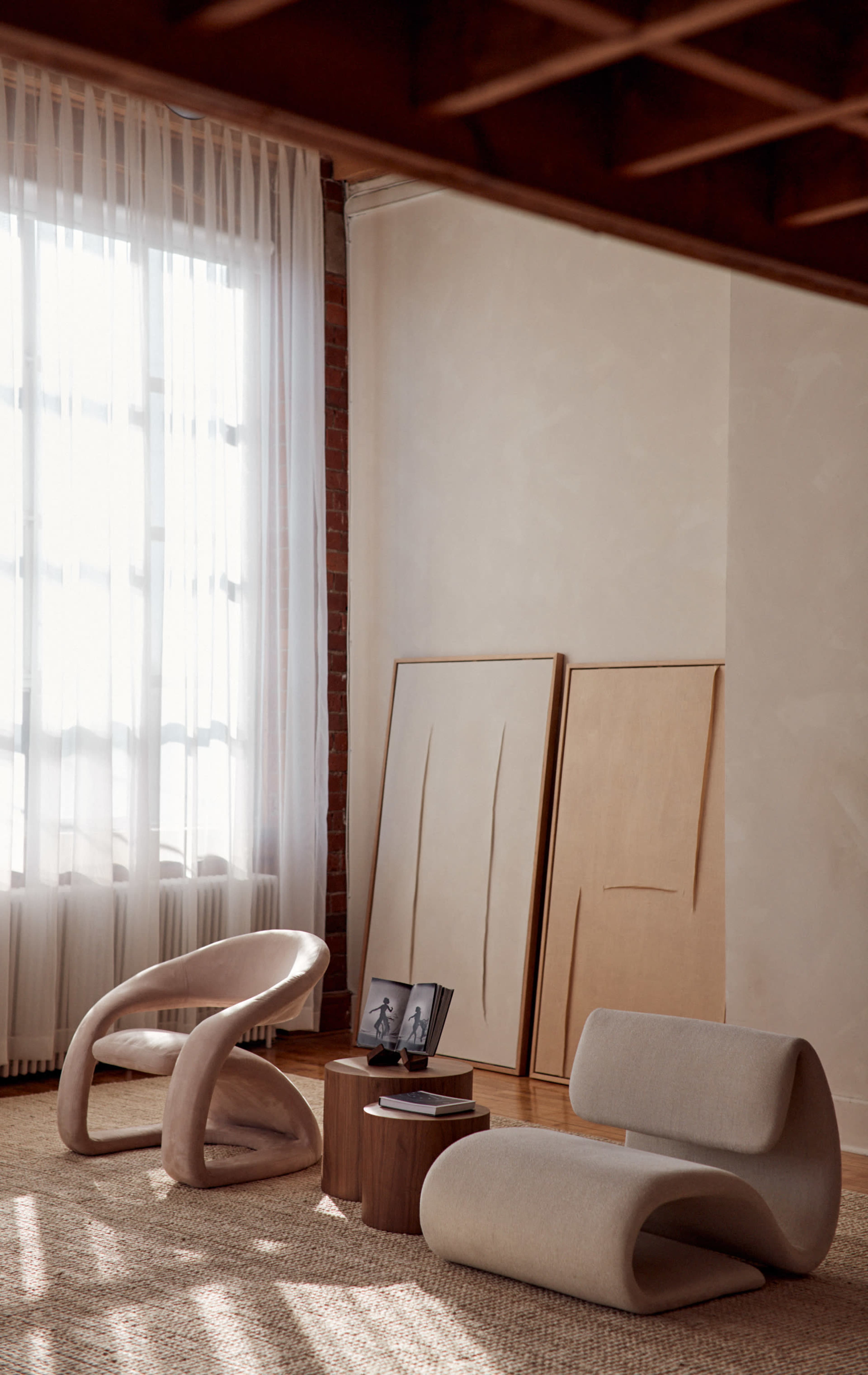 The image shows a stylish living area with two modern chairs, a wooden coffee table, and large windows dressed with sheer curtains, along with artwork leaning against the wall.