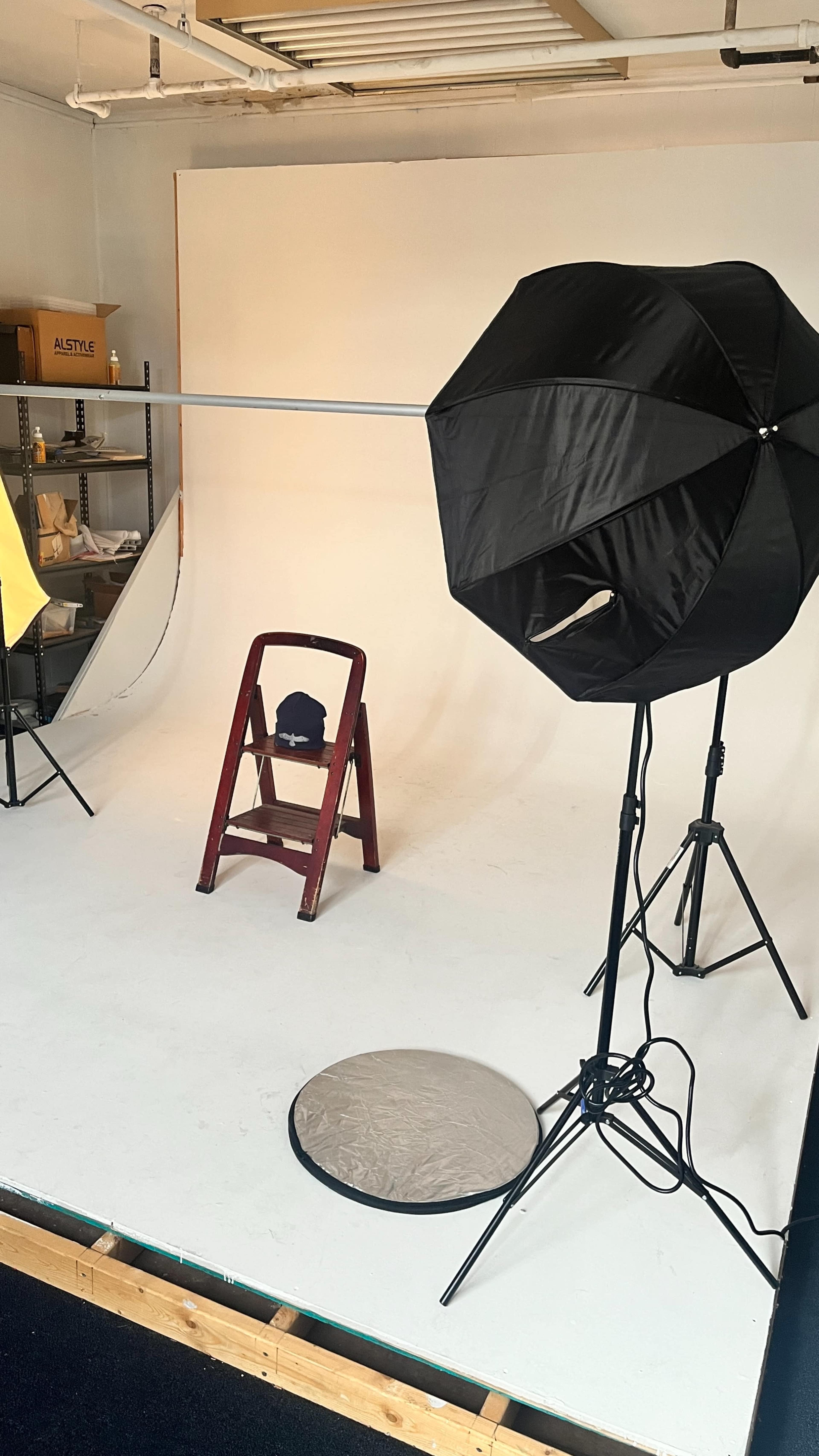 A photography studio setup, featuring a black umbrella light, a silver reflector, a wooden ladder, and a backdrop.