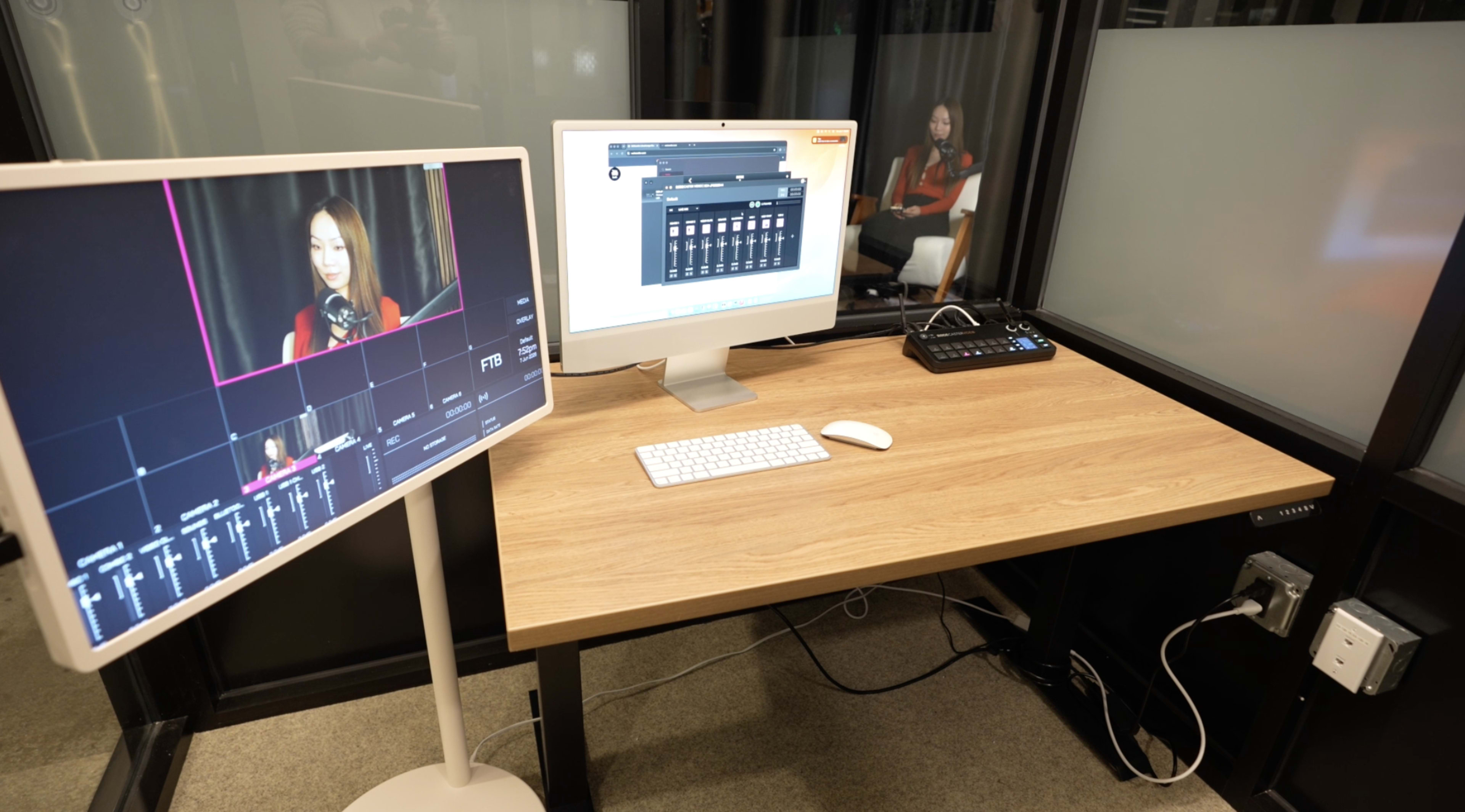 The image shows a modern workspace featuring two computer monitors, a keyboard, and a control panel, all set on a wooden desk in a glass-walled room.