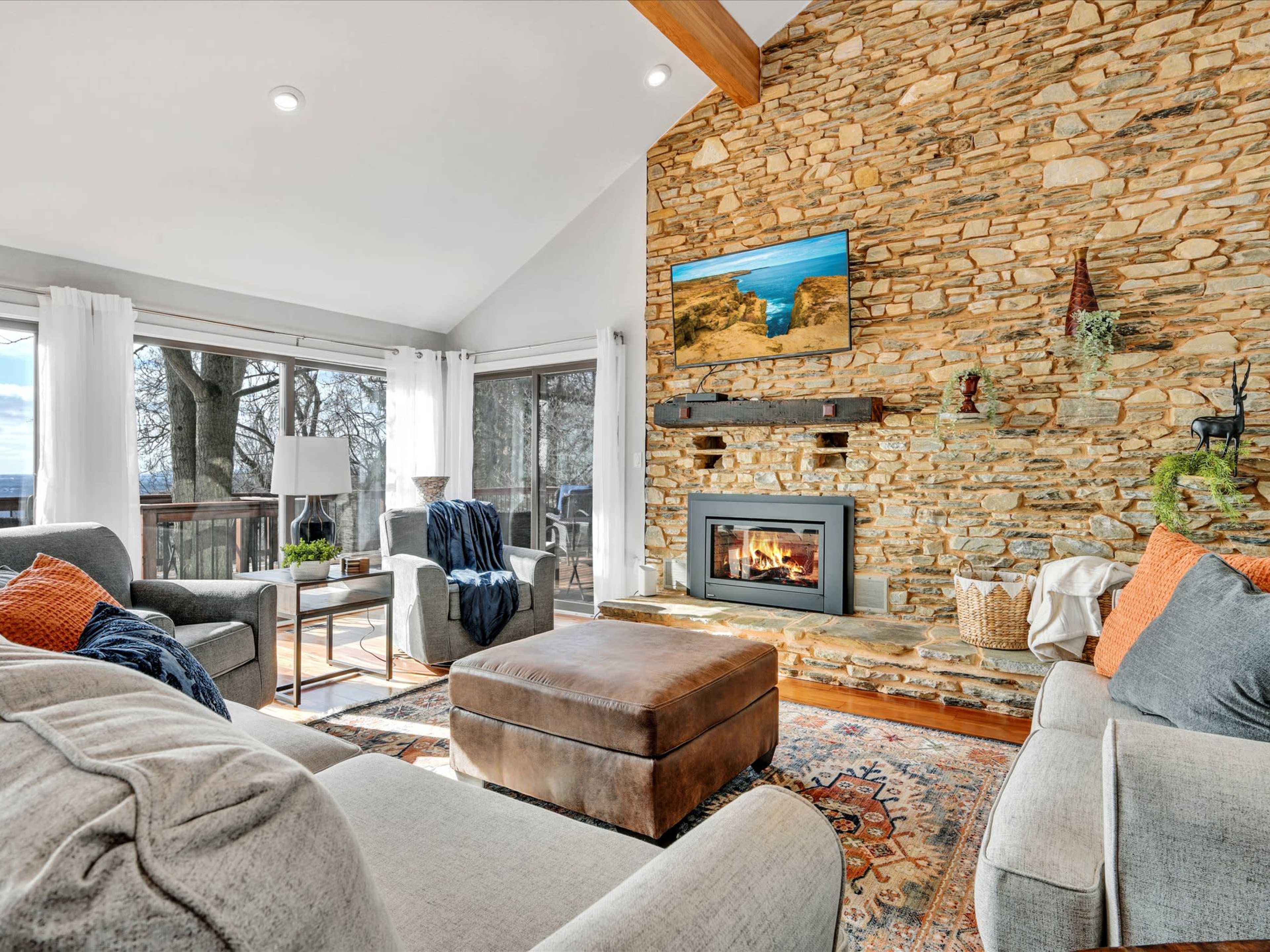 The living room features a stone fireplace, comfortable seating, and large windows overlooking a tree-filled view.