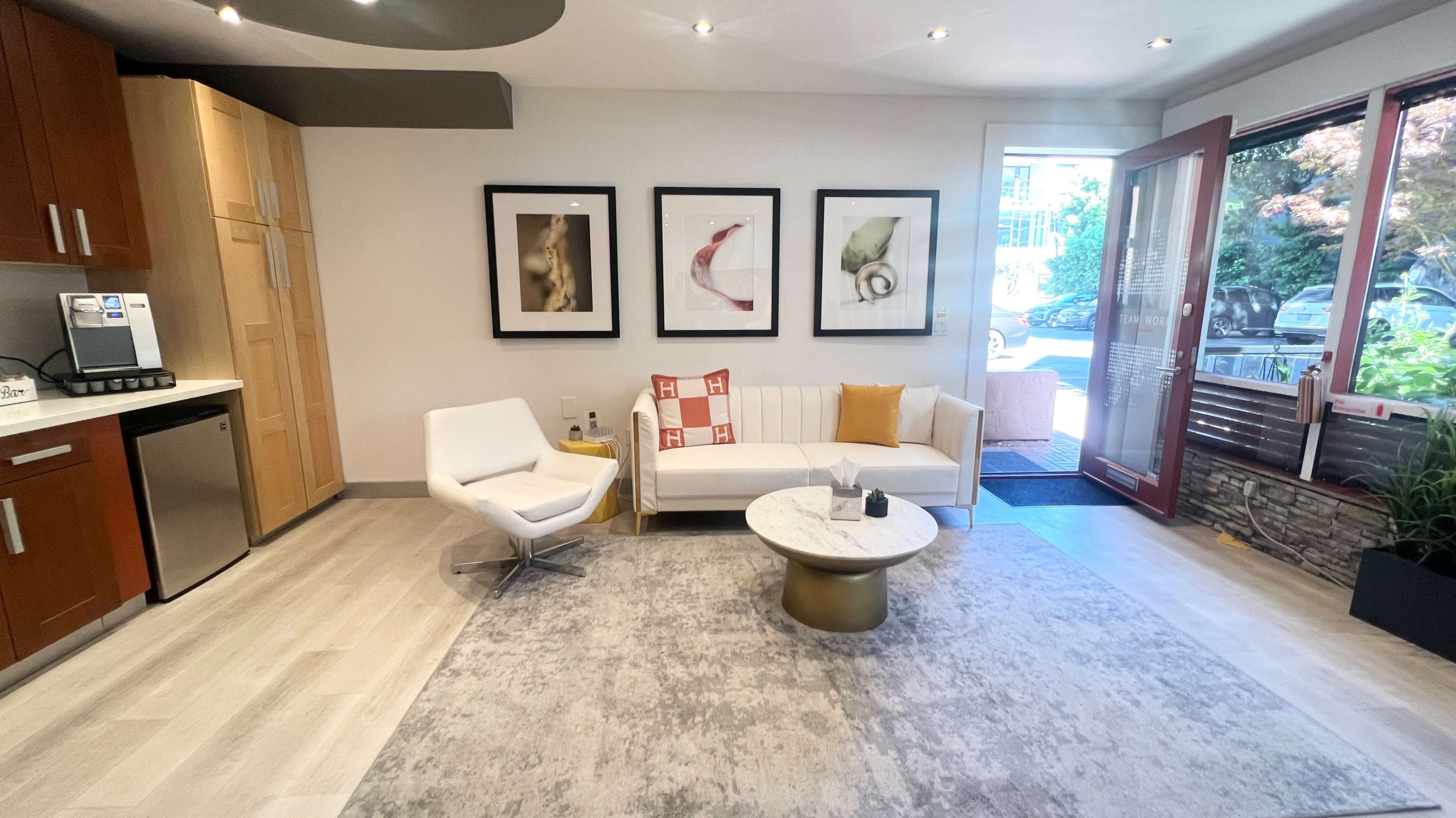 A modern living area with a white sofa, a round coffee table, and framed artwork on the walls, illuminated by natural light from a large window.