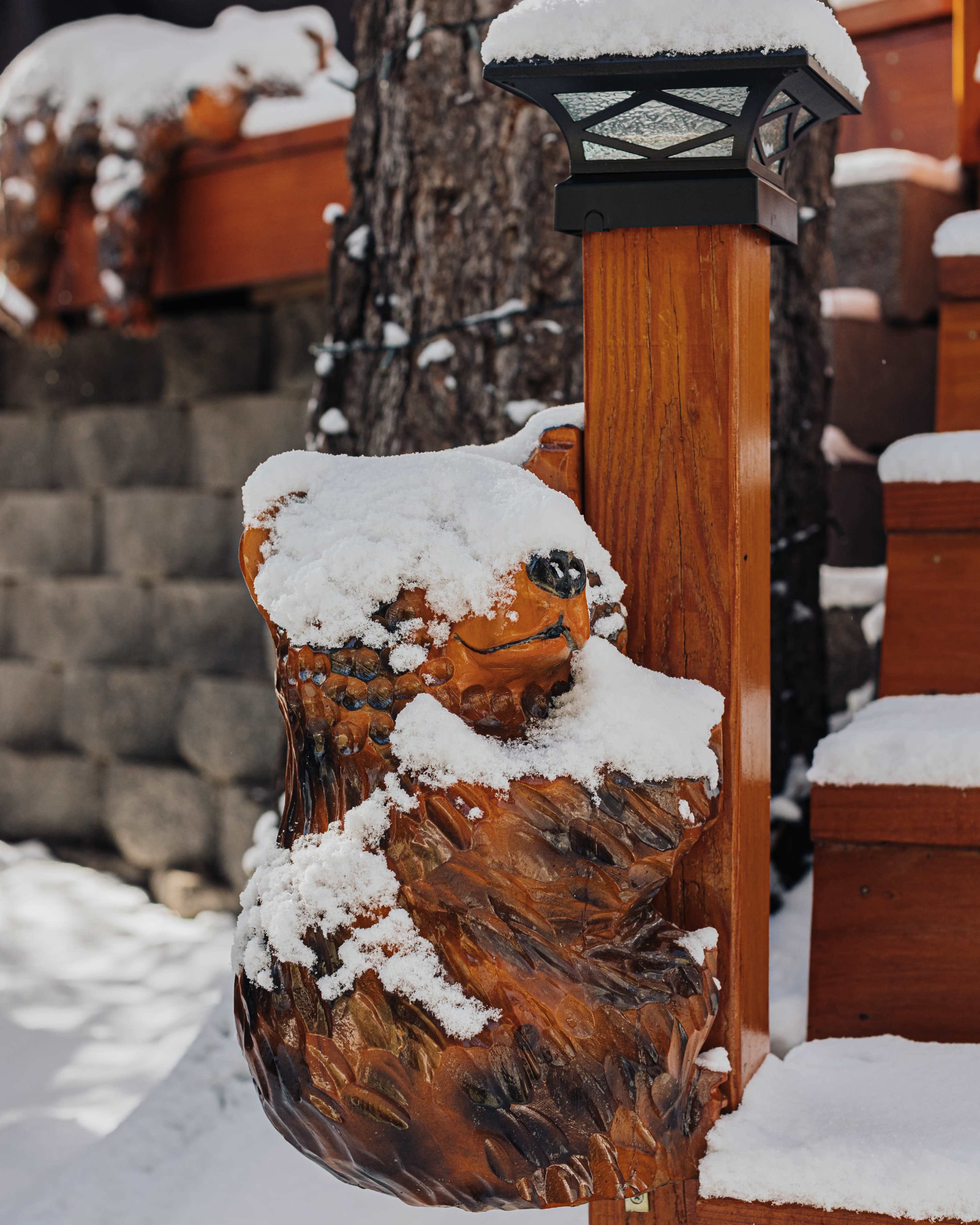 A wooden bear sculpture partially covered in snow sits on a wooden post beside a staircase.